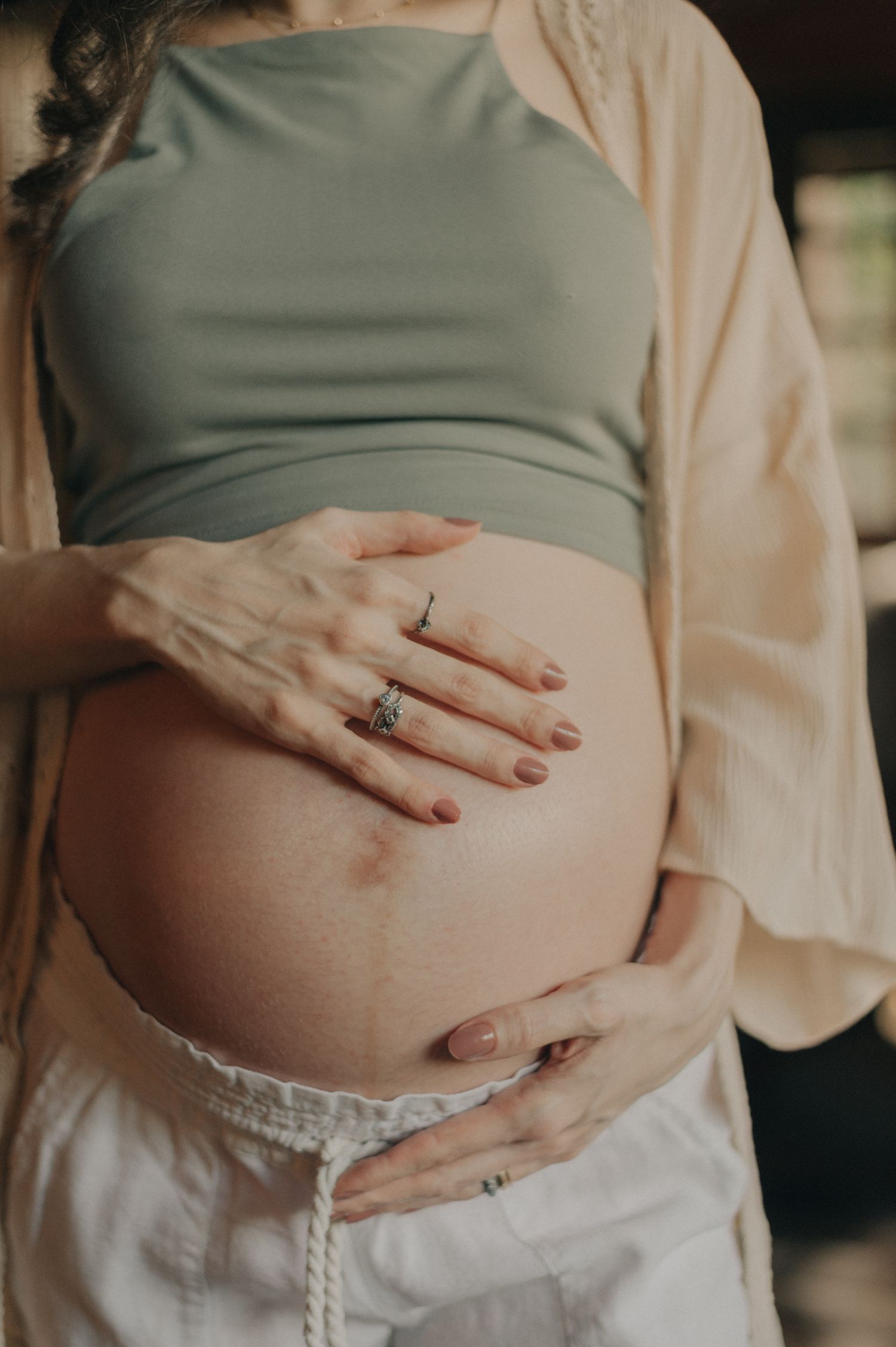 Close-up of hands with rings resting on pregnant belly in soft natural lighting.