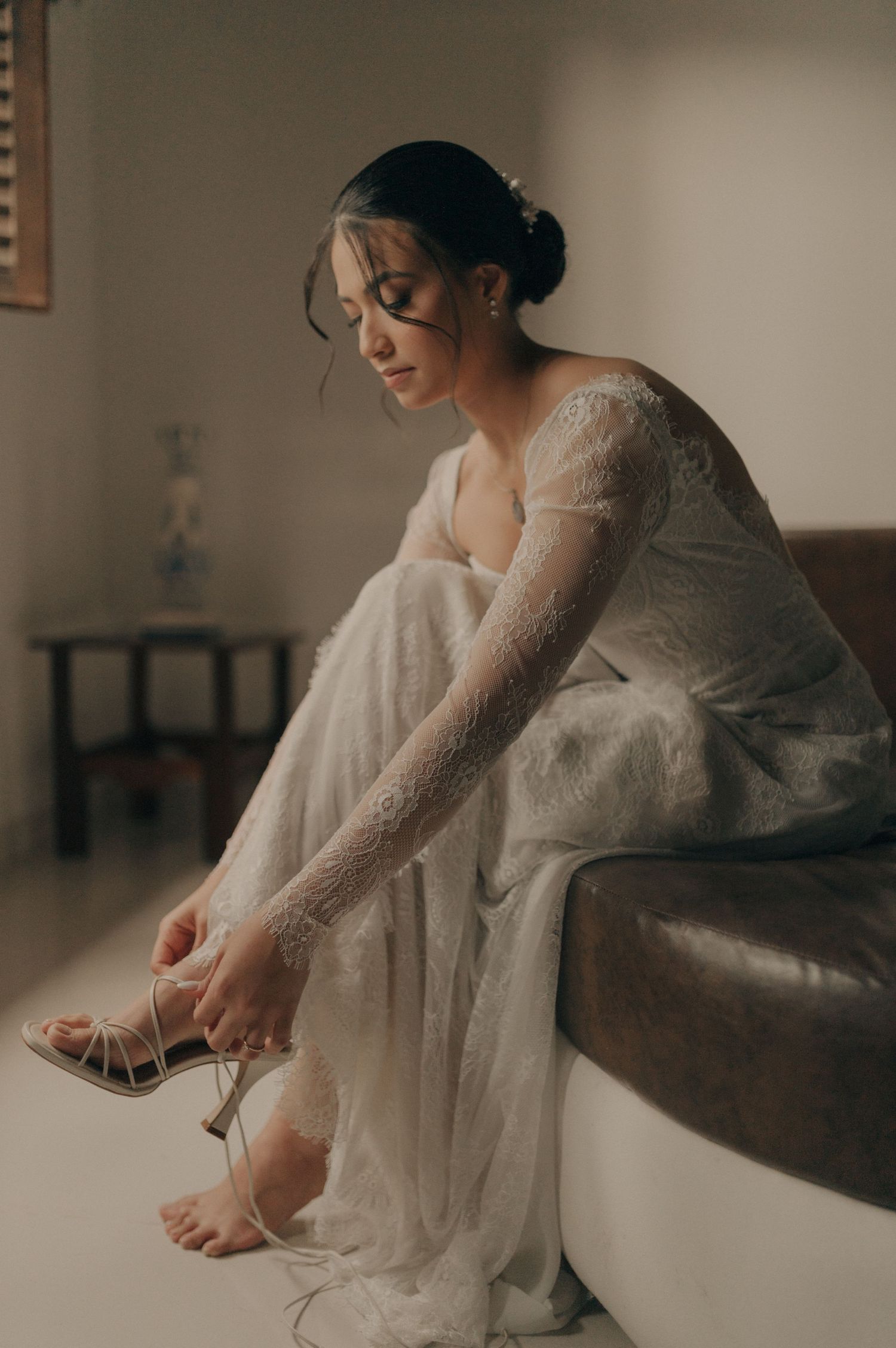 Bride in lace dress putting on her wedding shoes while sitting on a leather bench.