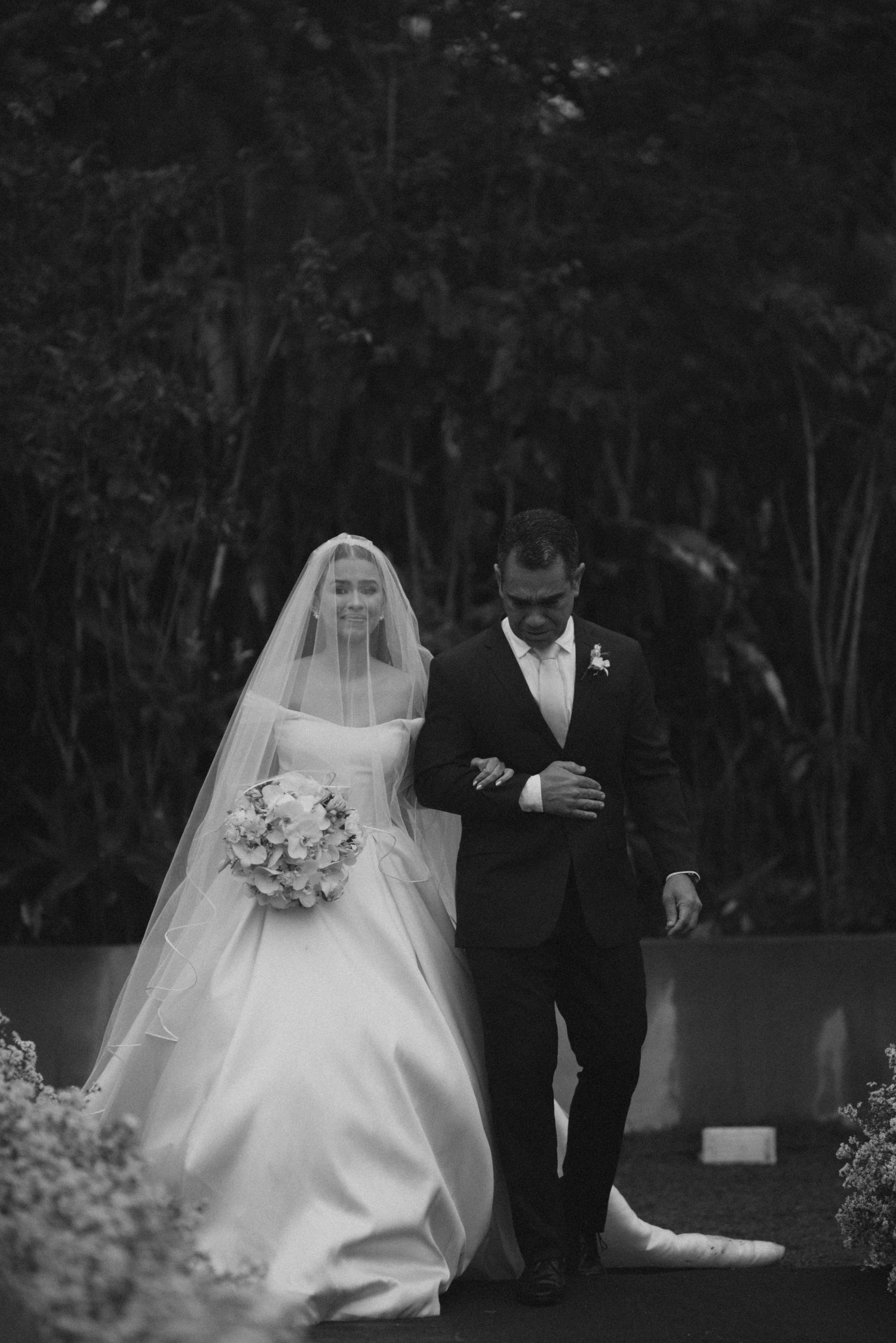 A black and white wedding photograph showing a couple walking together, with the bride in a flowing white gown and veil.