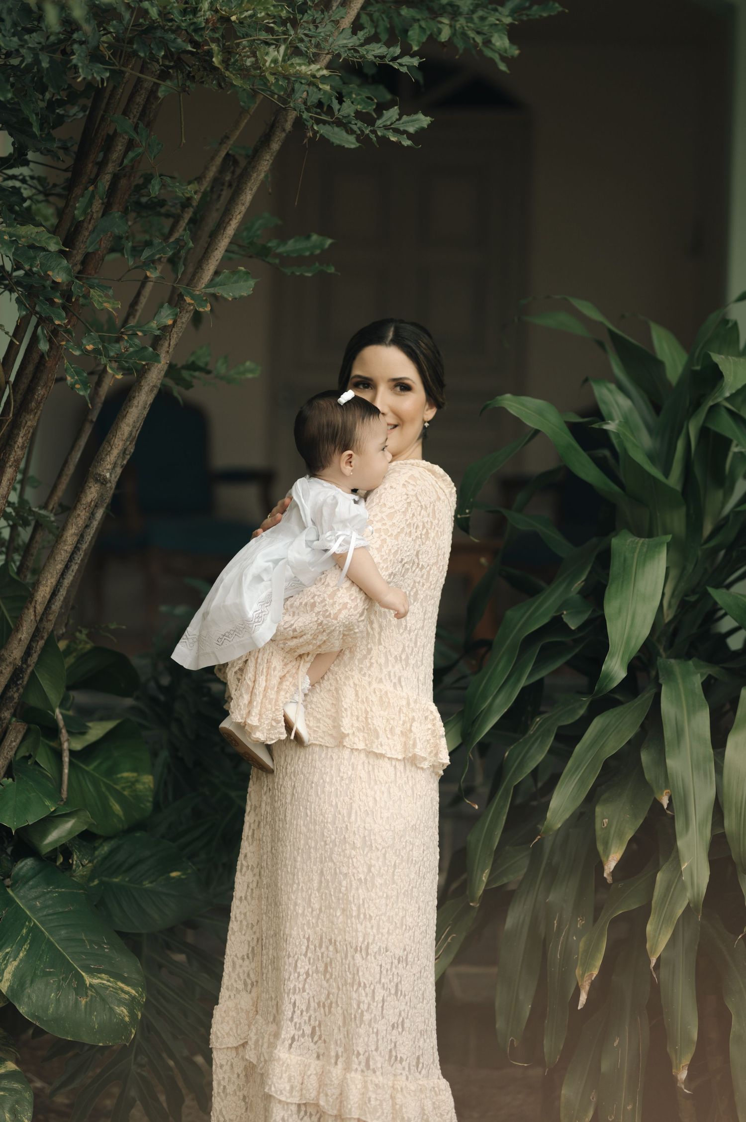 A tender moment captured in a garden setting with a parent wearing a delicate cream lace dress holding a baby in white.