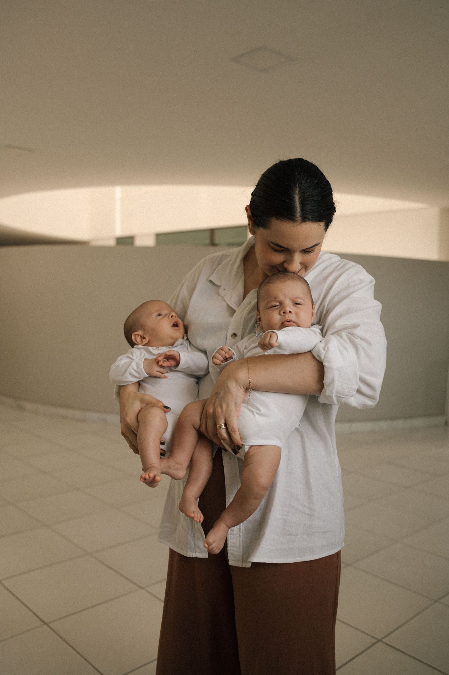 A tender moment between parent and baby in white and brown clothing against a minimalist interior setting.