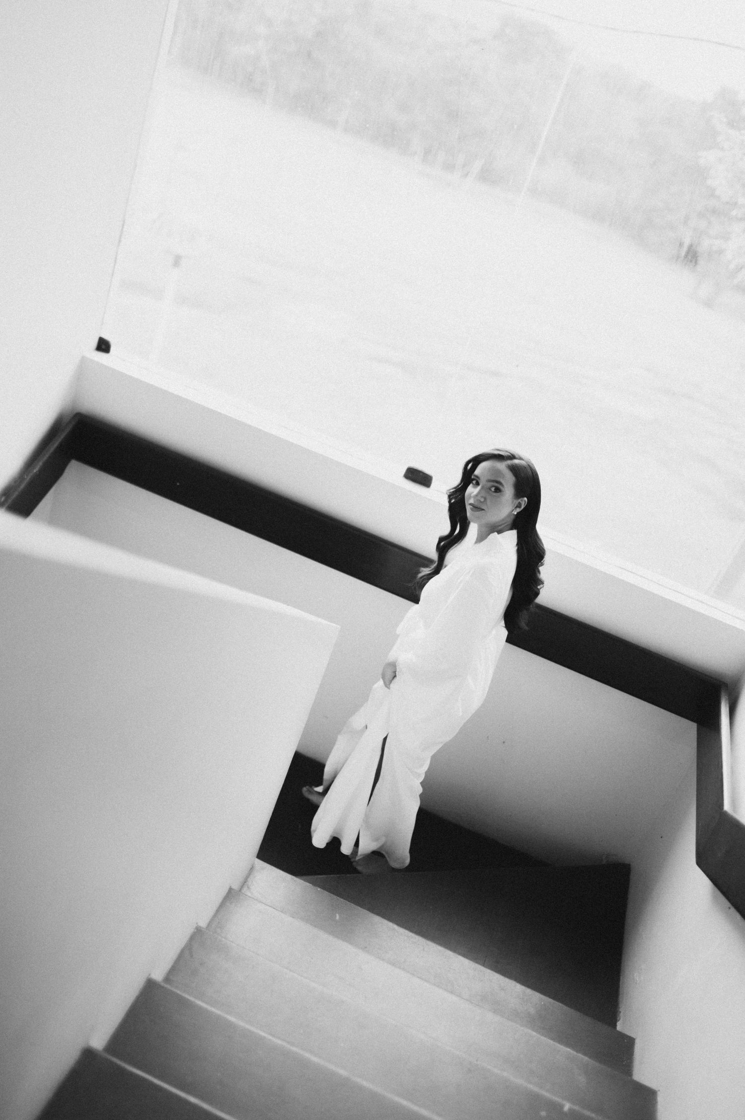 Black and white photo of a bride in a white dress standing on modern stairs, looking up at the camera.