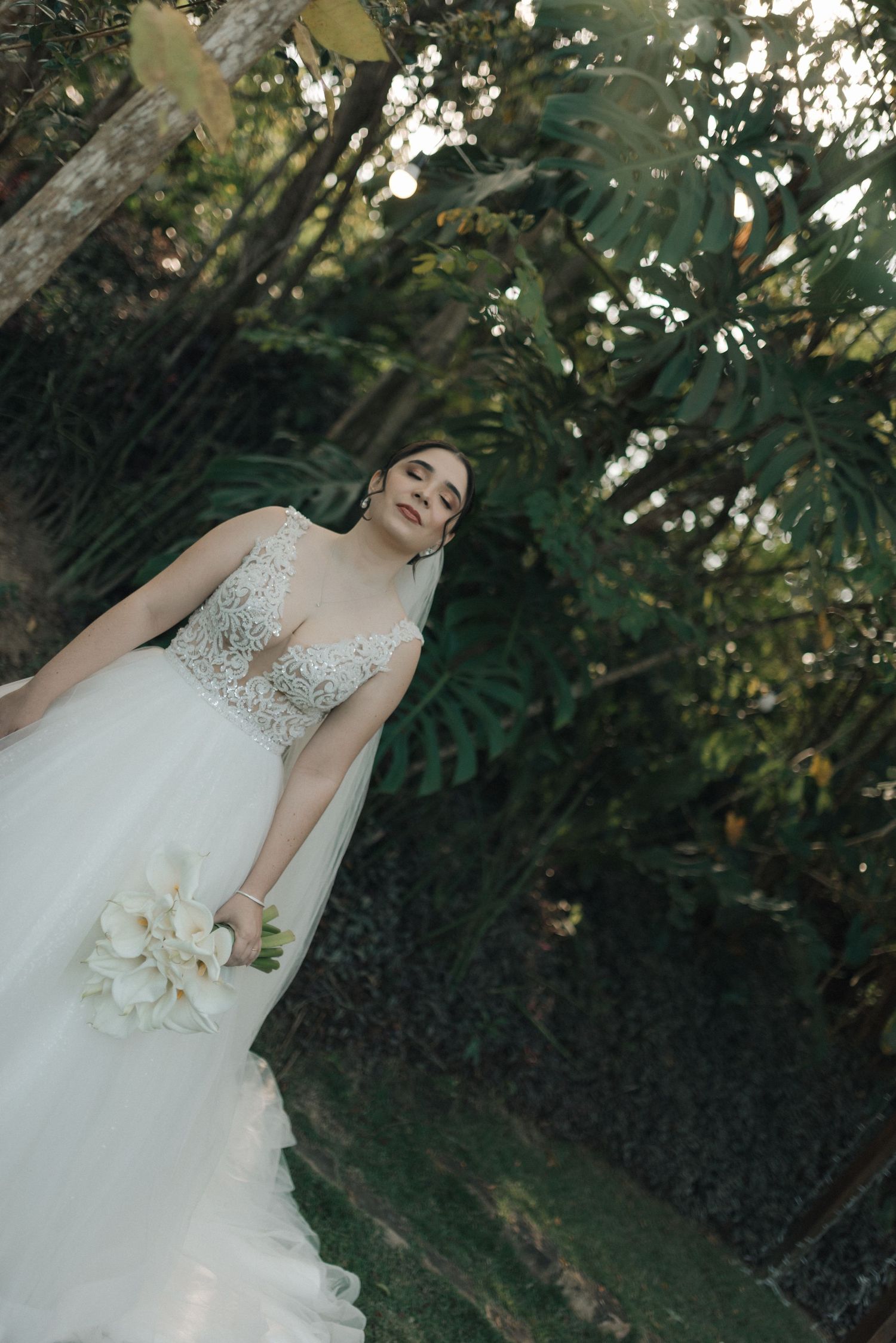 A bride in a lace wedding gown poses outdoors among garden greenery.