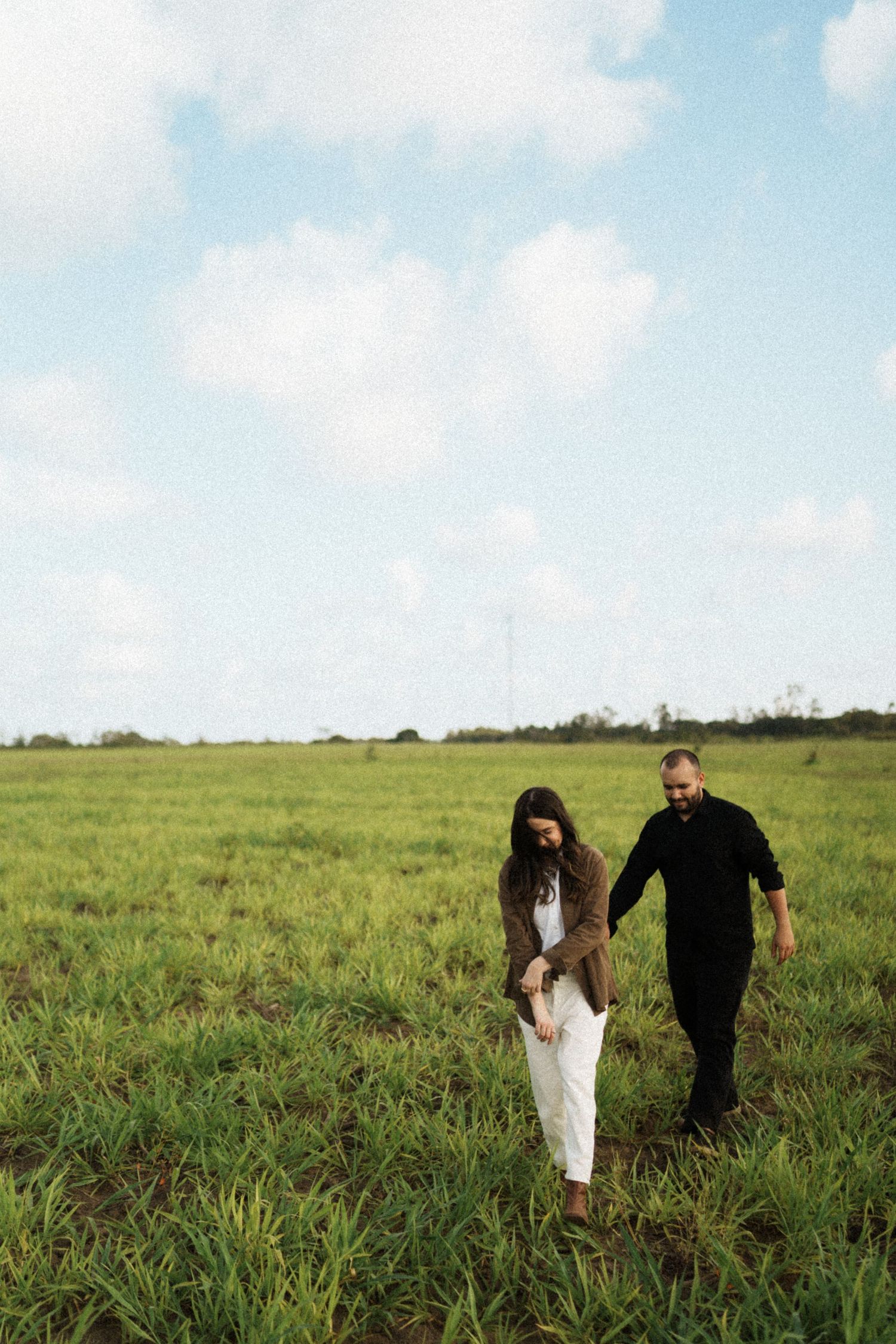 A couple walks through a lush green field under a bright blue sky with scattered clouds.