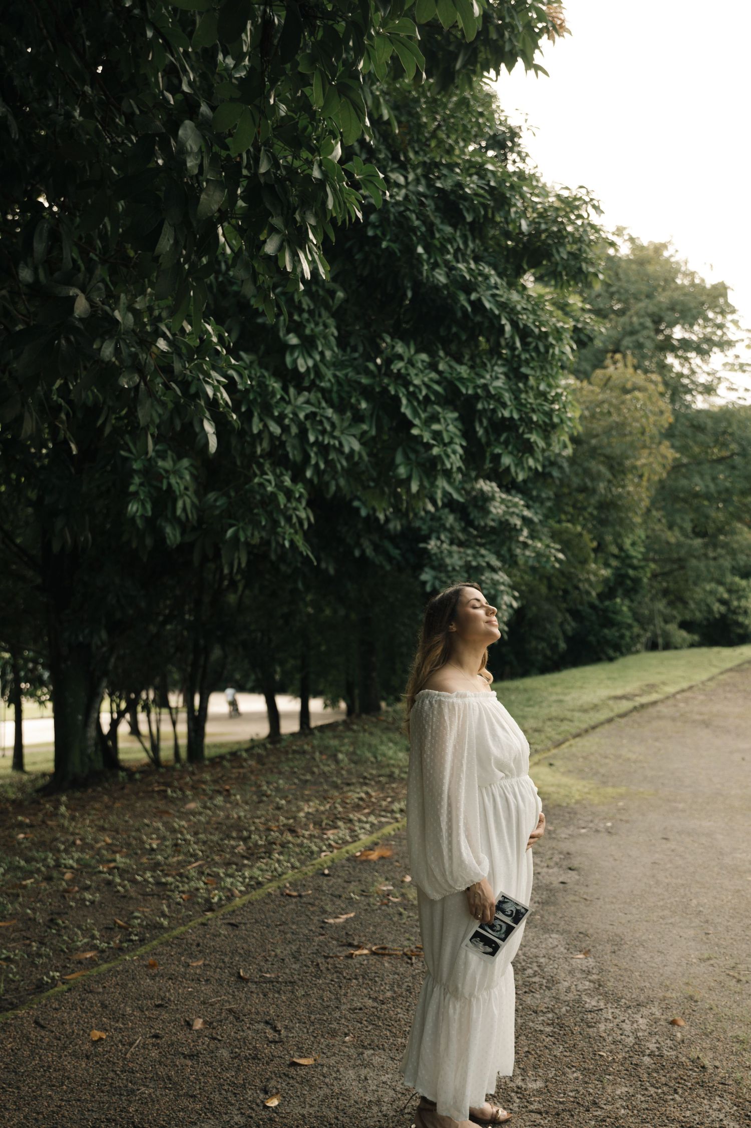 Pregnant woman in white off-shoulder dress holding ultrasound, standing in lush green park path, looking up serenely.