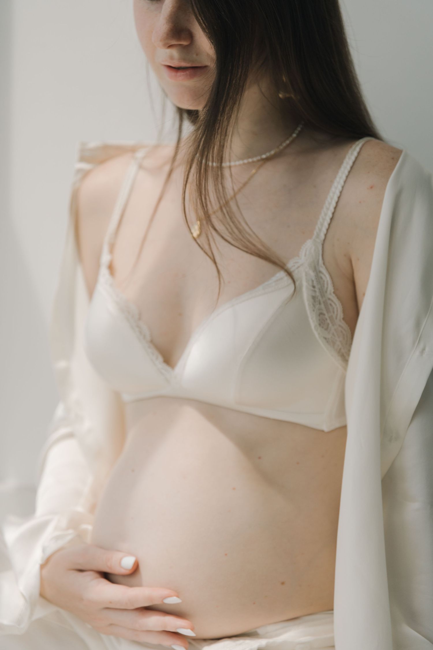 Soft and intimate maternity portrait in white lingerie with delicate jewelry details.
