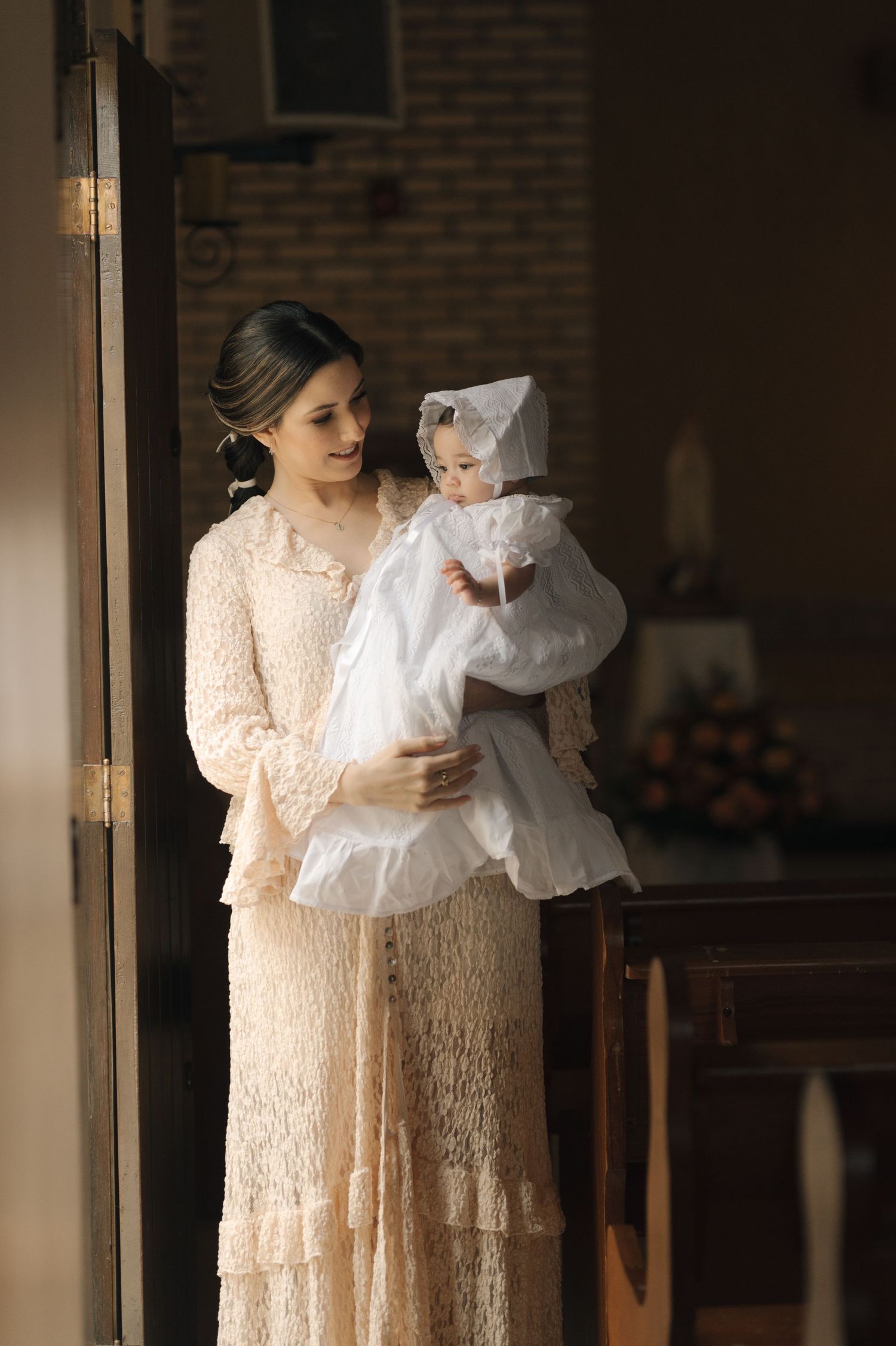 Mother in lace dress holds baby in traditional white baptismal gown near window with natural light.