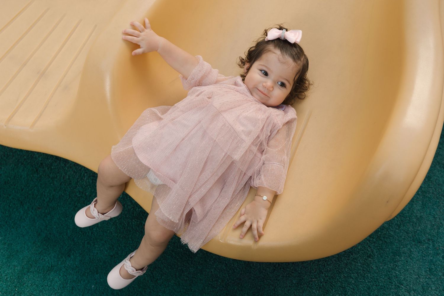 Happy toddler in pink tulle dress plays on yellow playground slide.