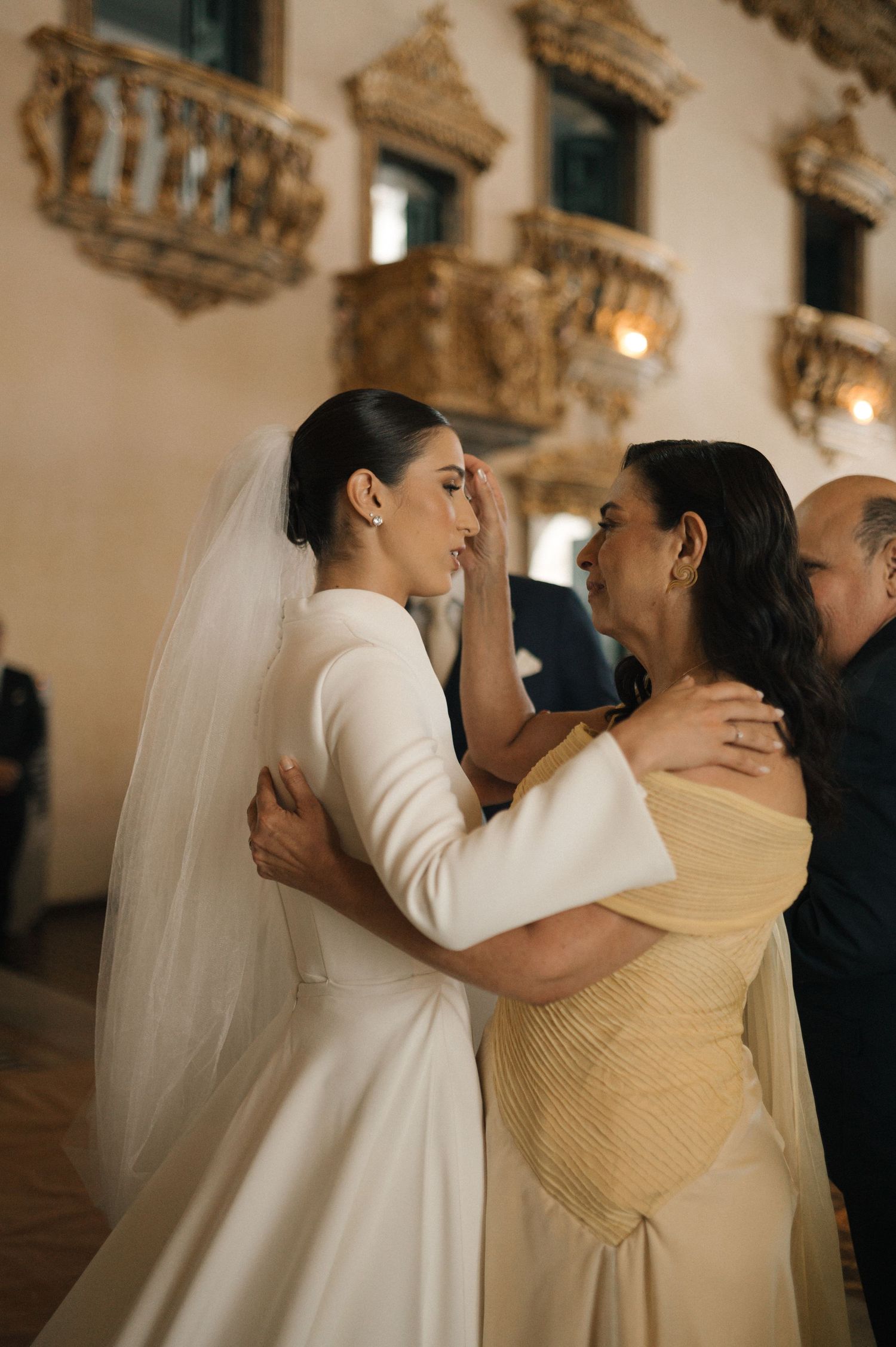Bride in white gown and veil shares emotional moment with mother in golden dress at elegant venue.