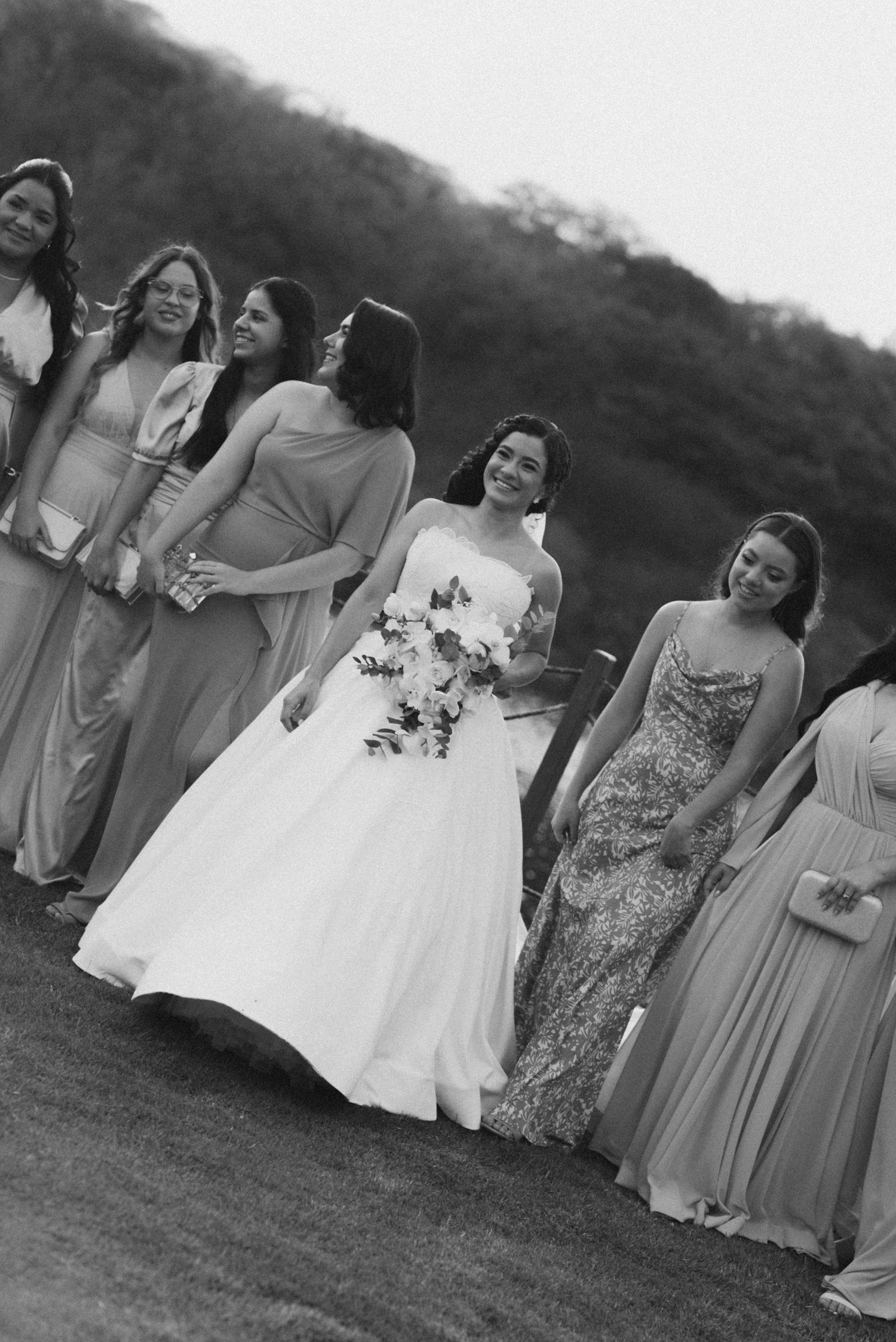 A bridal party in pastel dresses poses together on a grassy hillside.
