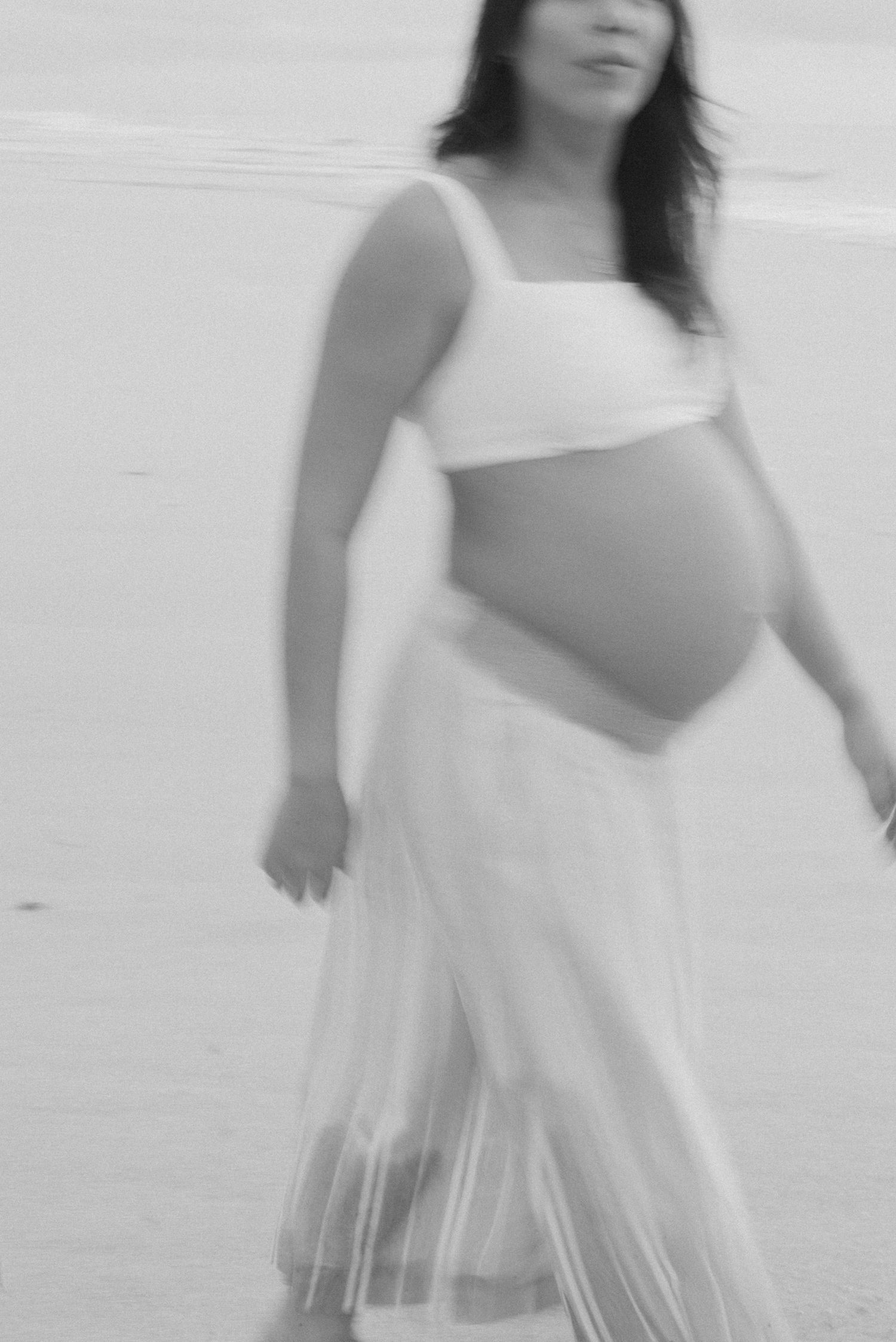 A soft black and white maternity photo showing an expectant mother in a flowing white dress.