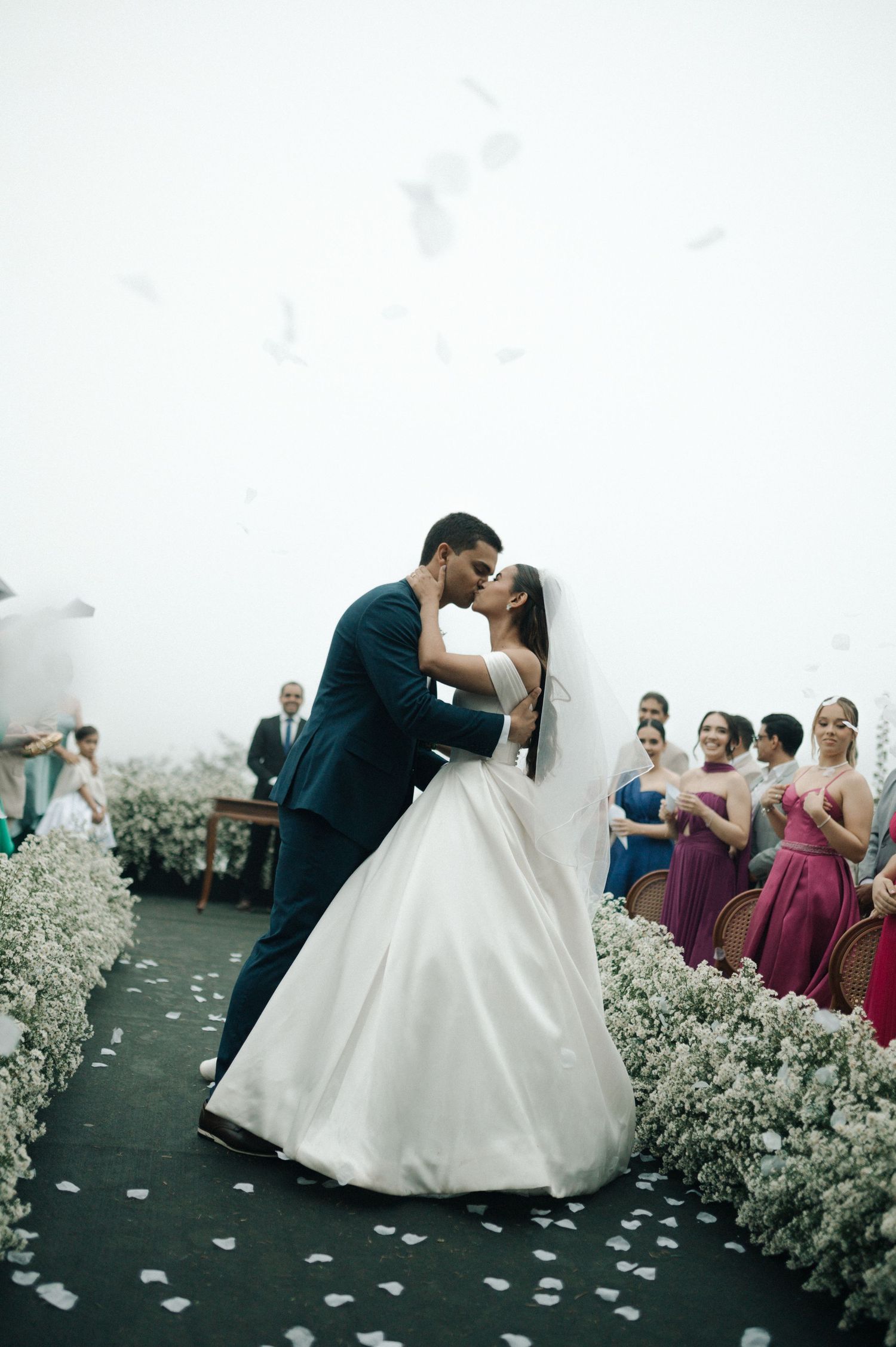 A romantic wedding couple shares their first dance surrounded by white floral decorations and rose petals on the floor.