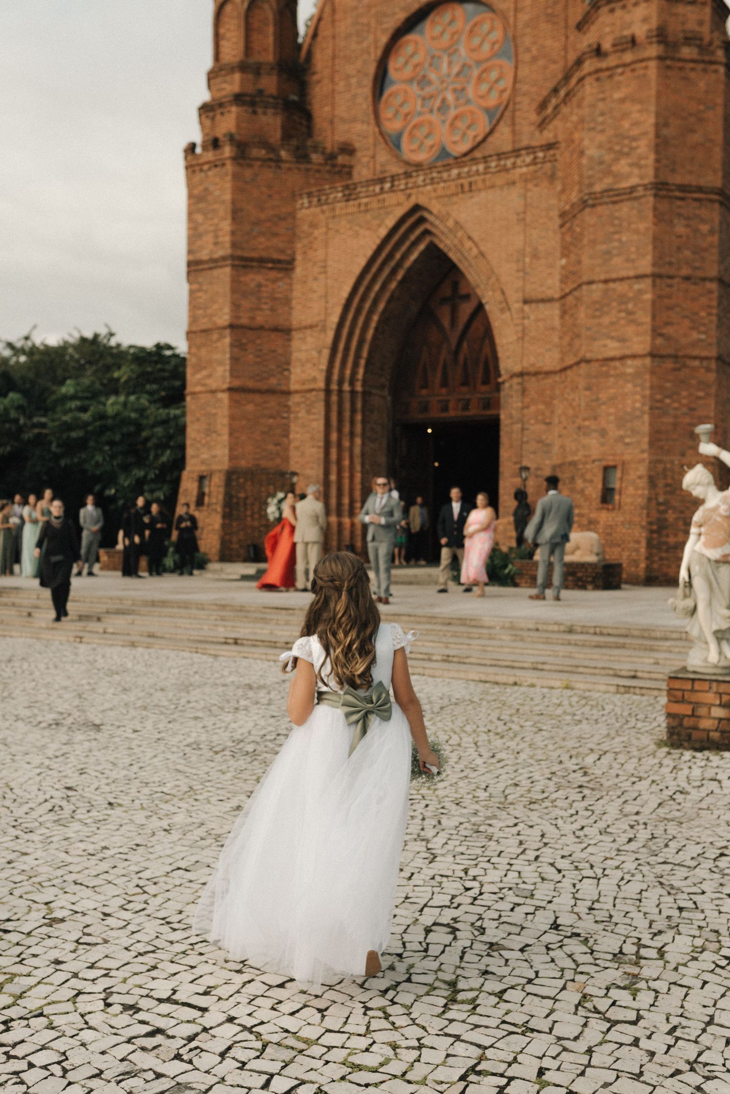 A bride in a white wedding dress stands on cobblestone steps in front of a historic brick church with gothic arches.