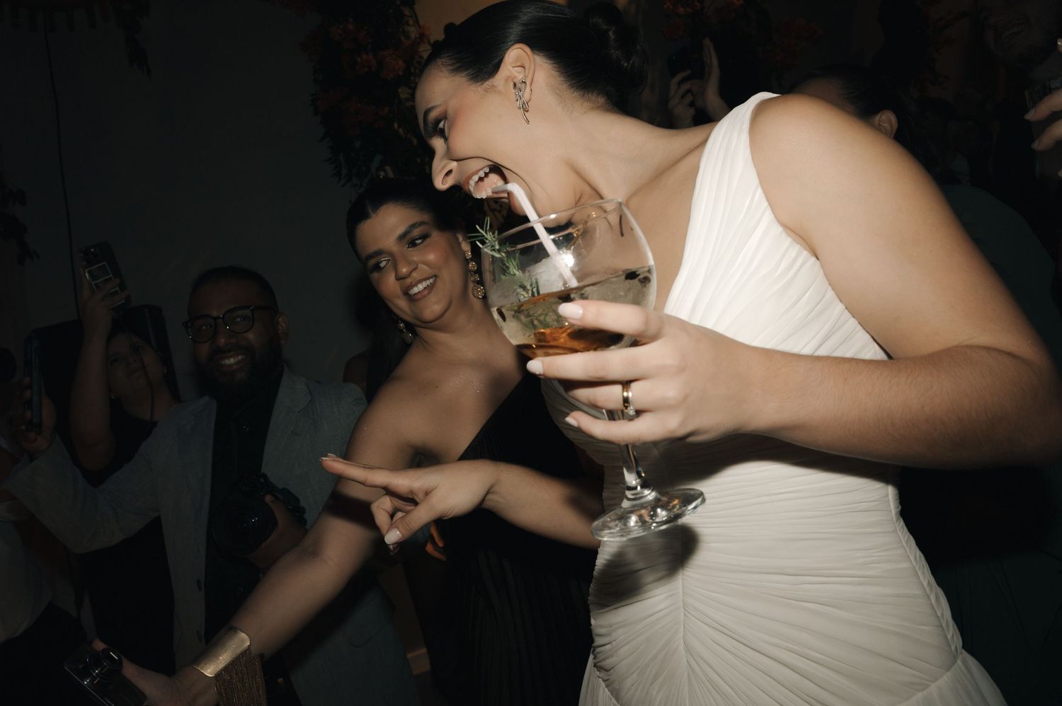 People enjoying drinks and dancing at a celebration party in dark lighting with someone wearing a white dress.