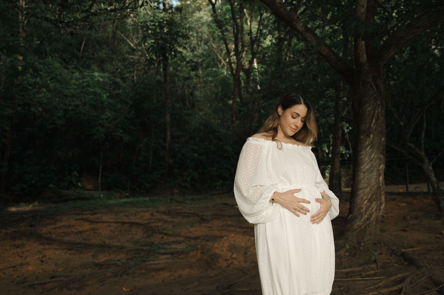 Pregnant woman in white dress cradling baby bump outdoors among trees during maternity photo shoot.
