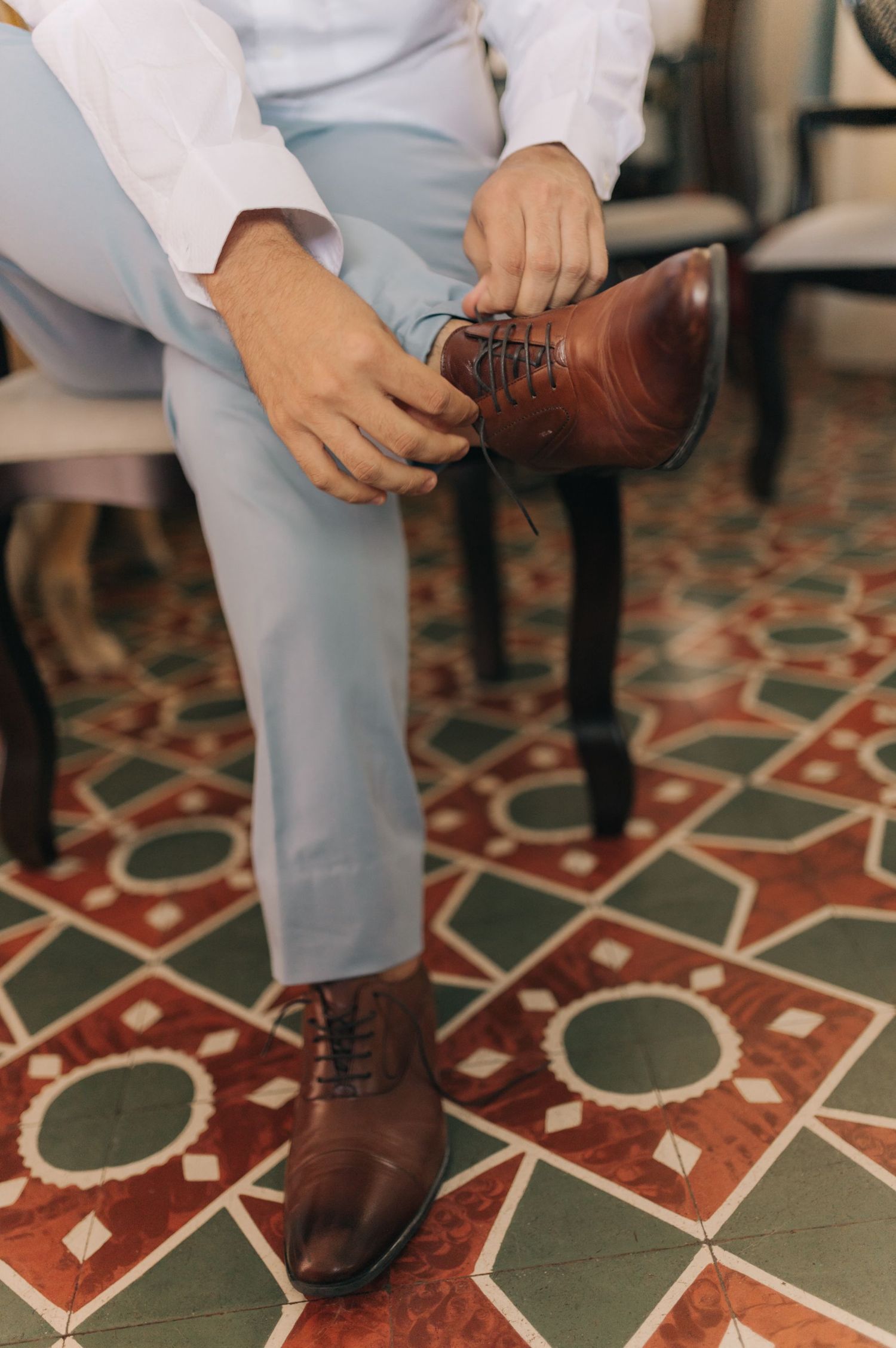 Close-up of hands tying brown leather dress shoes on tiled floor.