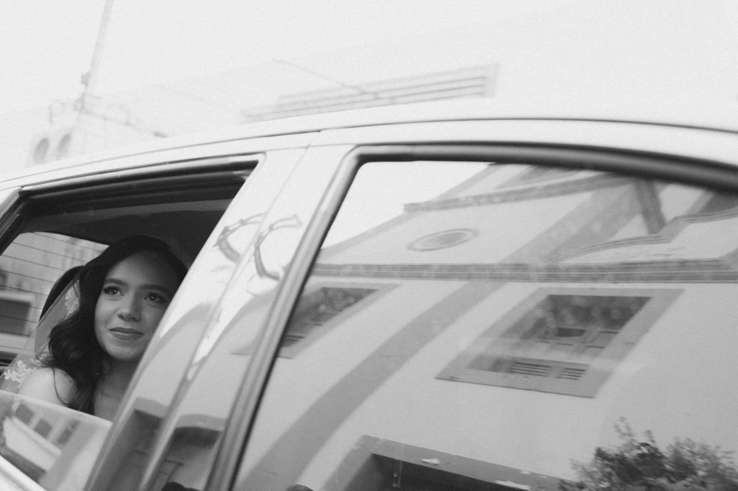 Black and white photo of a woman wearing a hat, gazing out of a car window with building reflections.