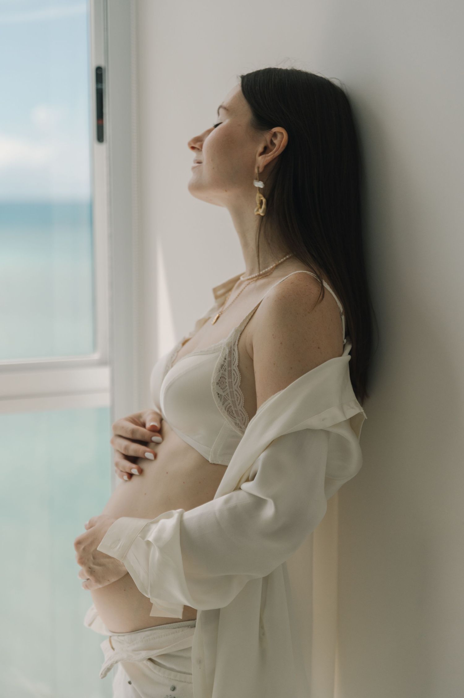 Serene maternity portrait by window in white lace lingerie and flowing robe.
