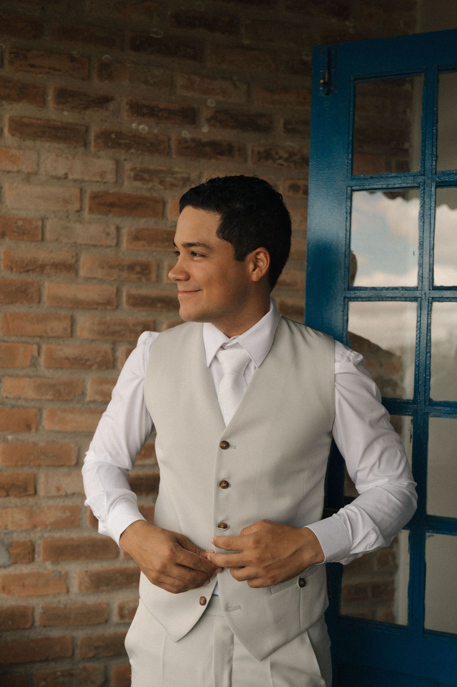A person in a white dress shirt and gray vest adjusts their clothing while standing near a brick wall with blue window frames.