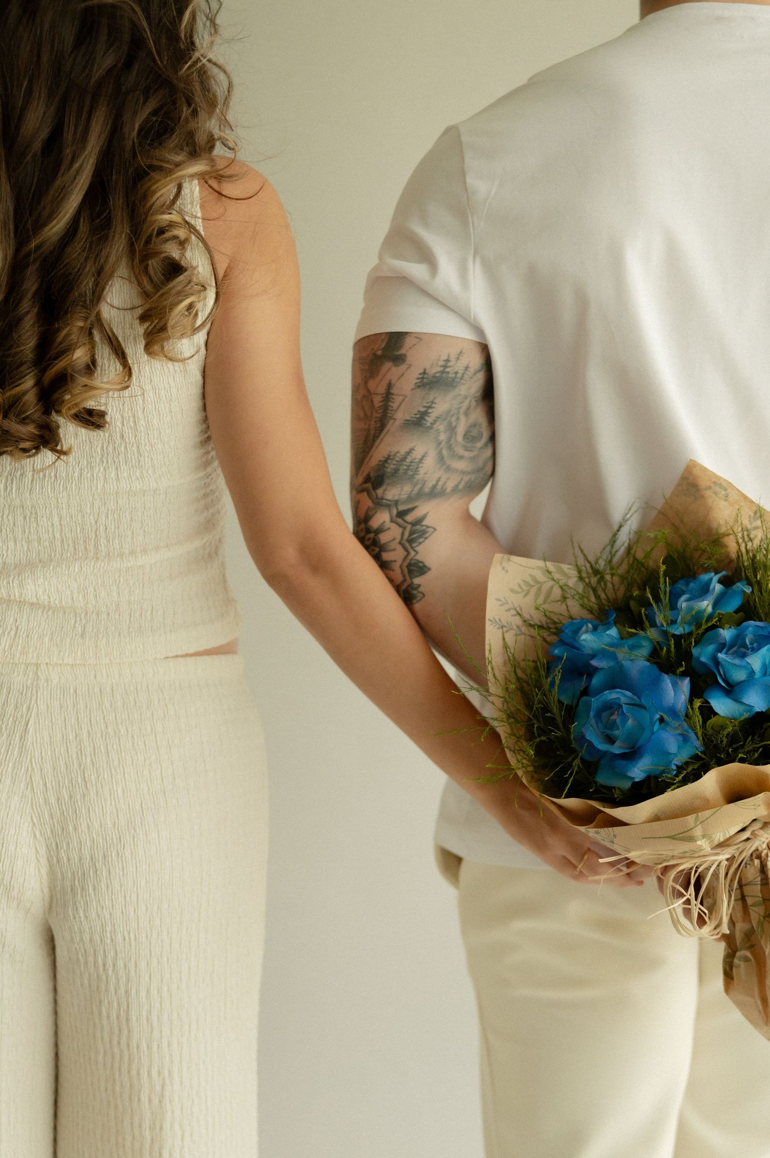 Couple in white outfits sharing a bouquet of blue roses and wheat, with visible tattoo on person's arm.