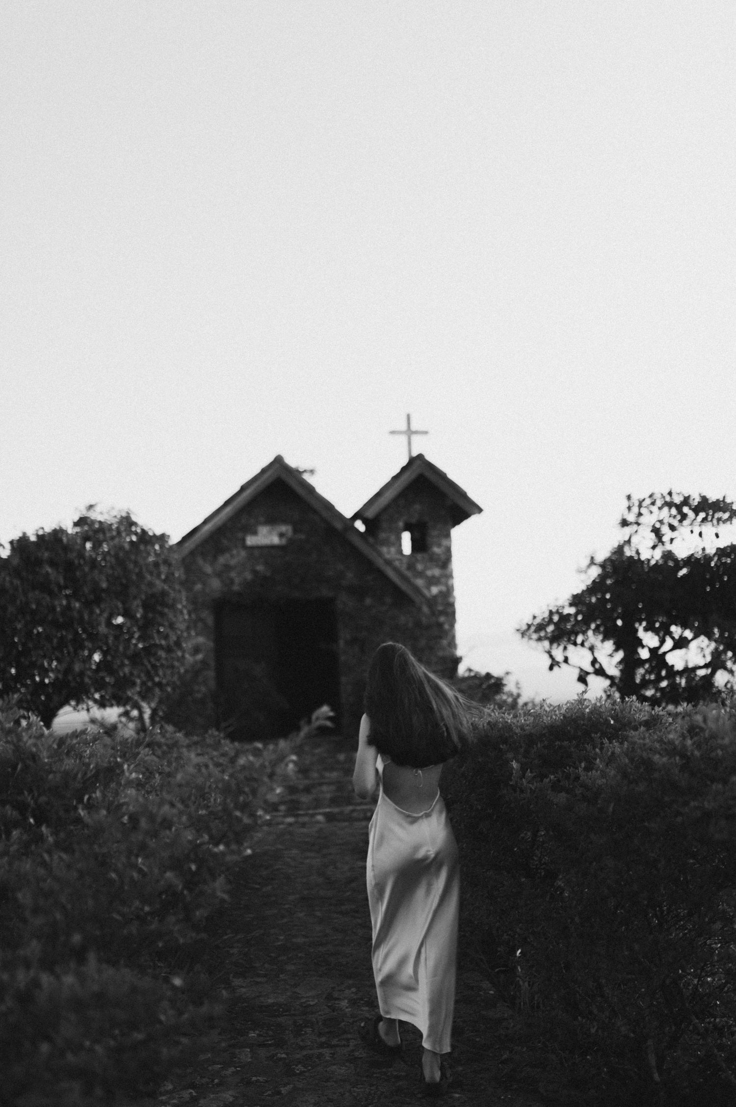 A figure in a white dress walks toward a small chapel on a hill in black and white.