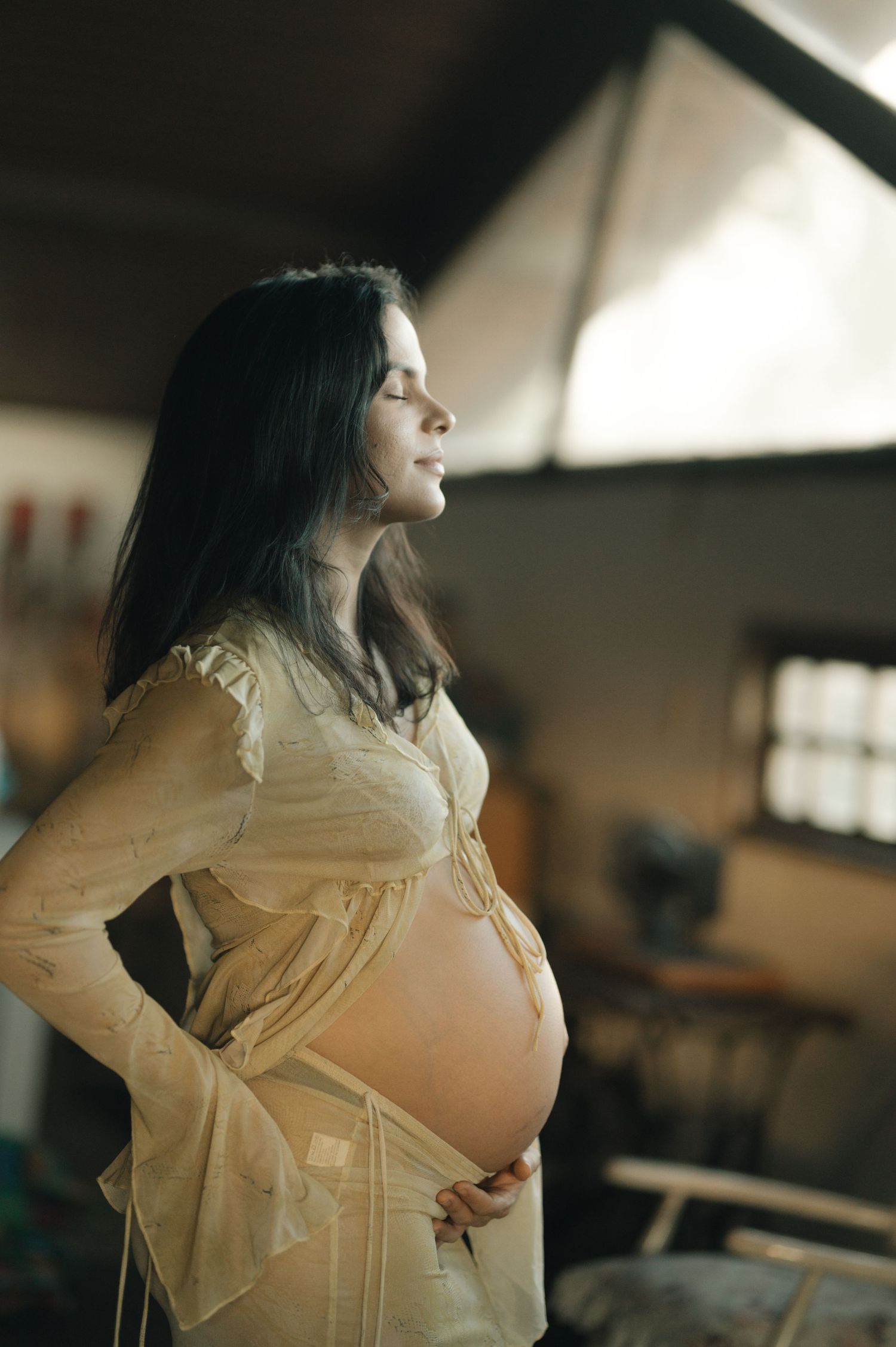 Expectant mother in white lace dress poses in profile by window light during maternity photoshoot.