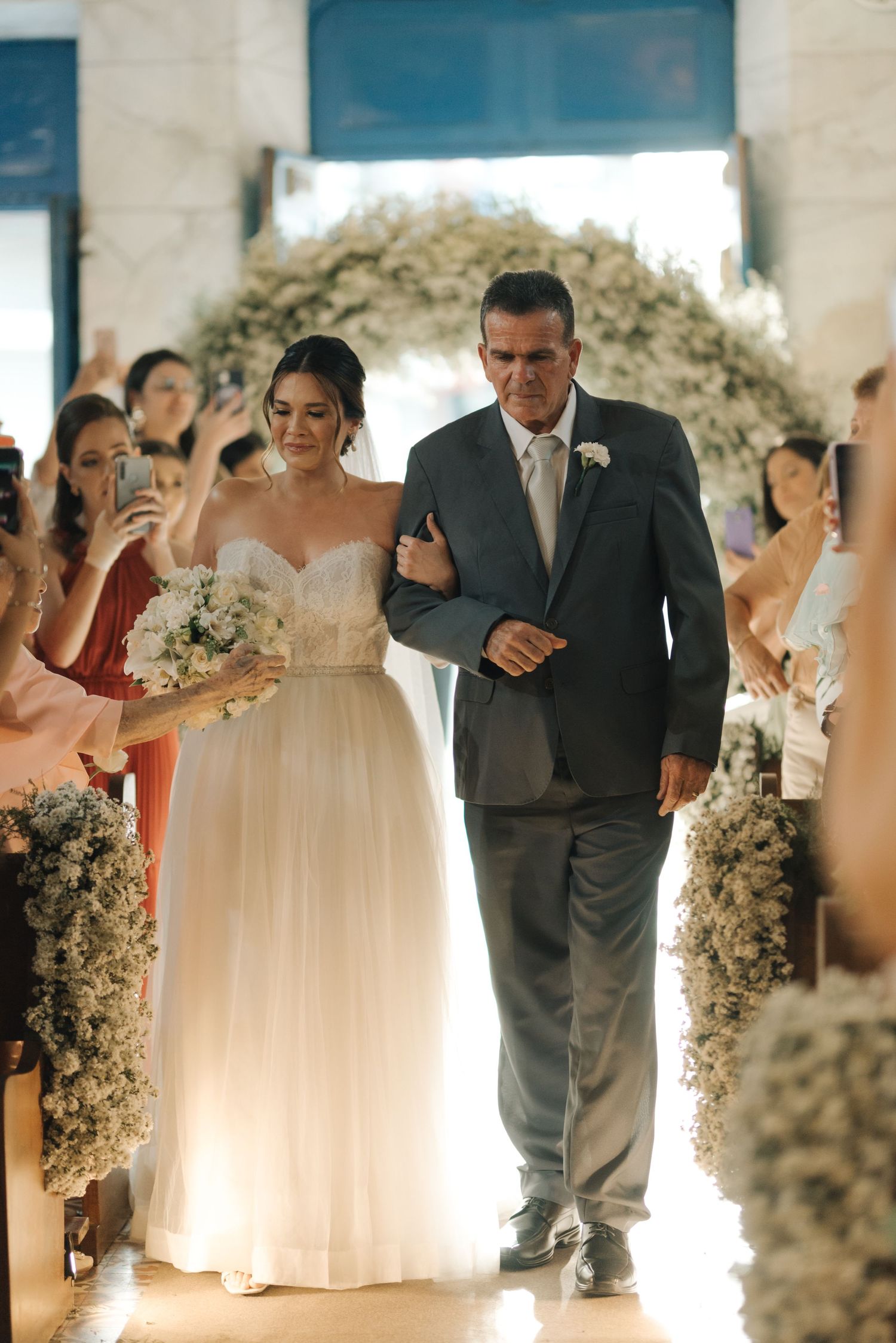 A bride in white gown walks down the aisle with her father under a floral arch.