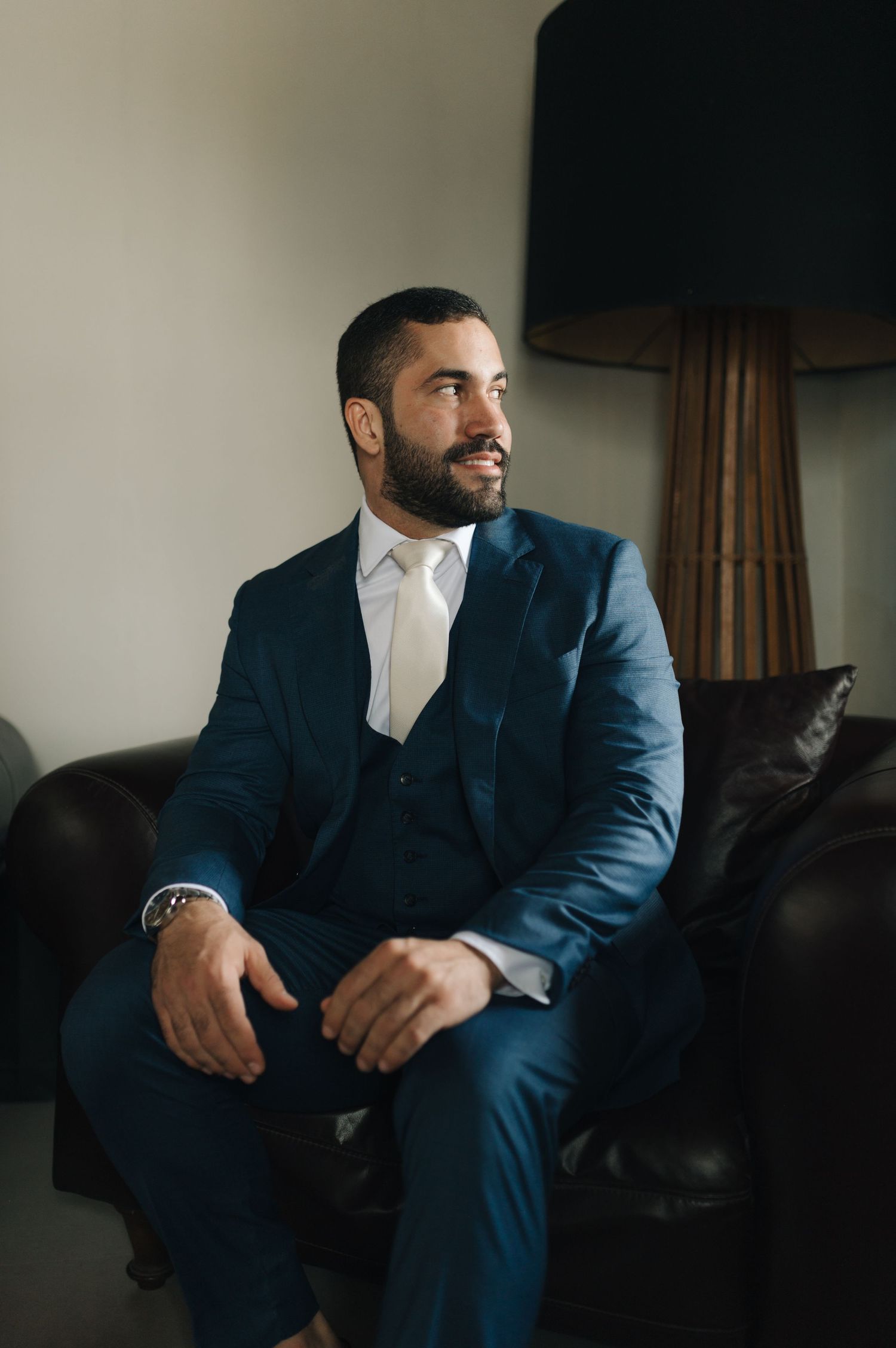 Groom in a tailored navy three-piece suit sitting on a leather couch before the ceremony.