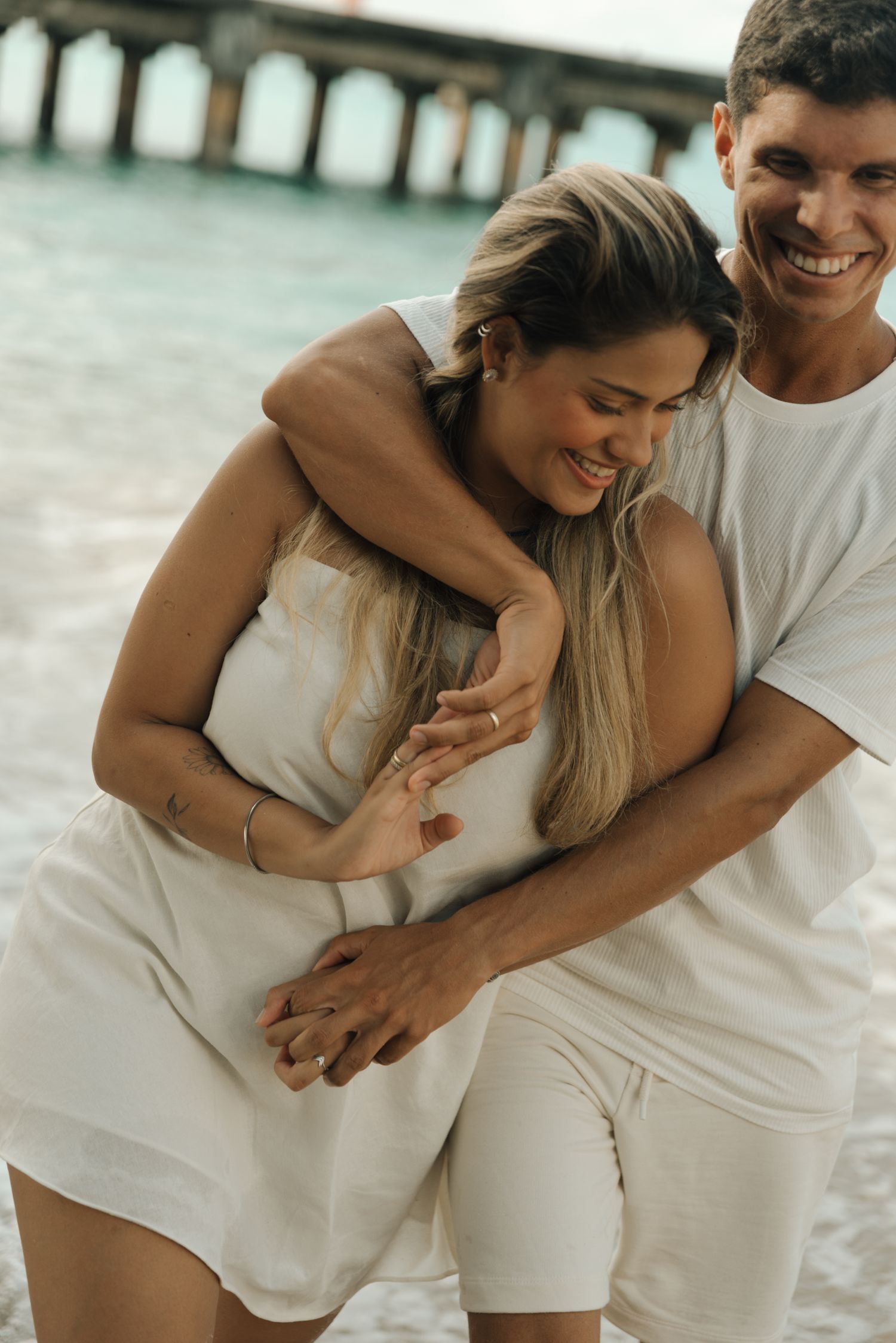 Joyful couple in white clothing laughing and embracing by the ocean.