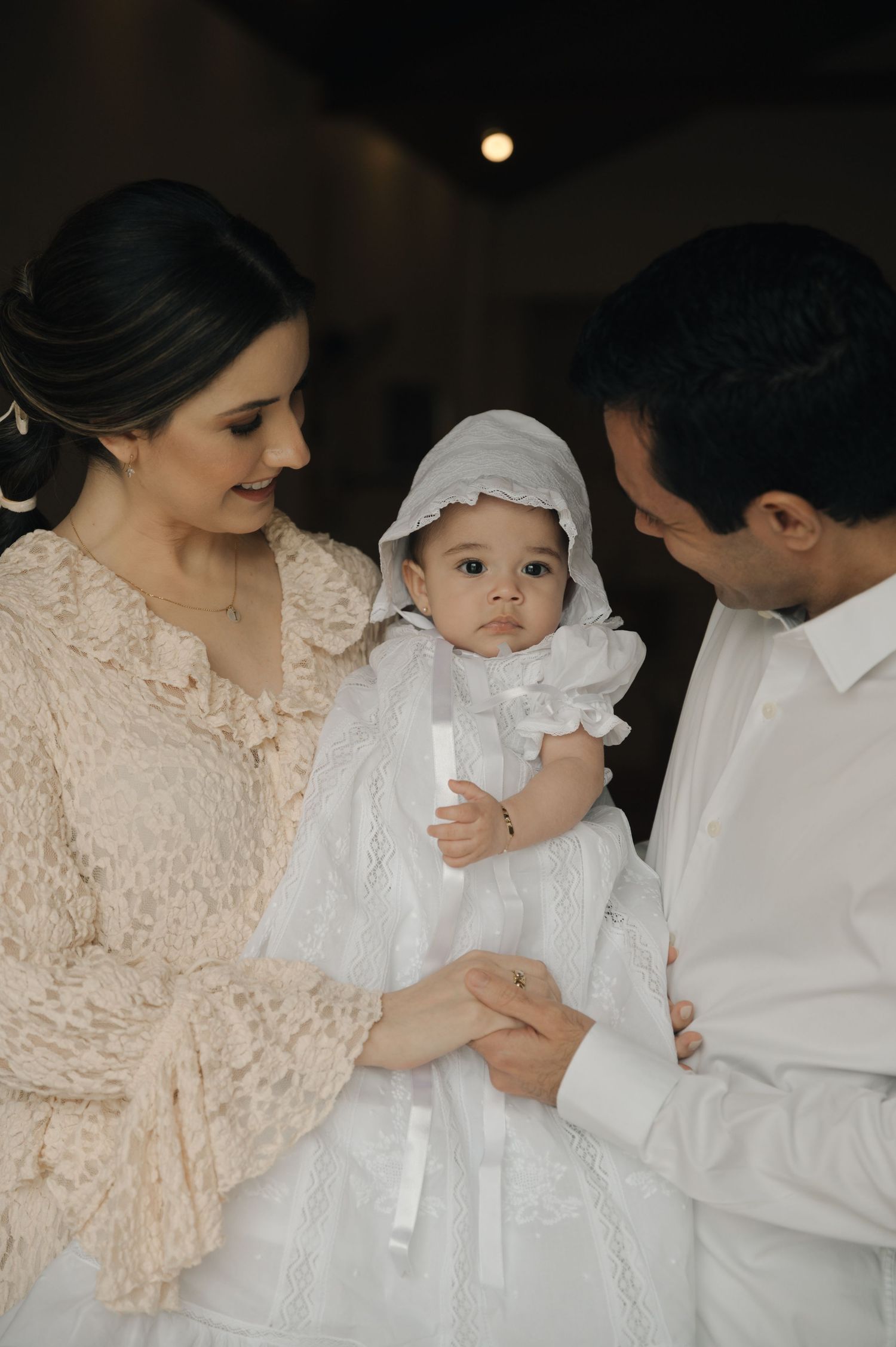 A tender family moment captured in soft lighting as parents hold their baby wearing a white christening gown.