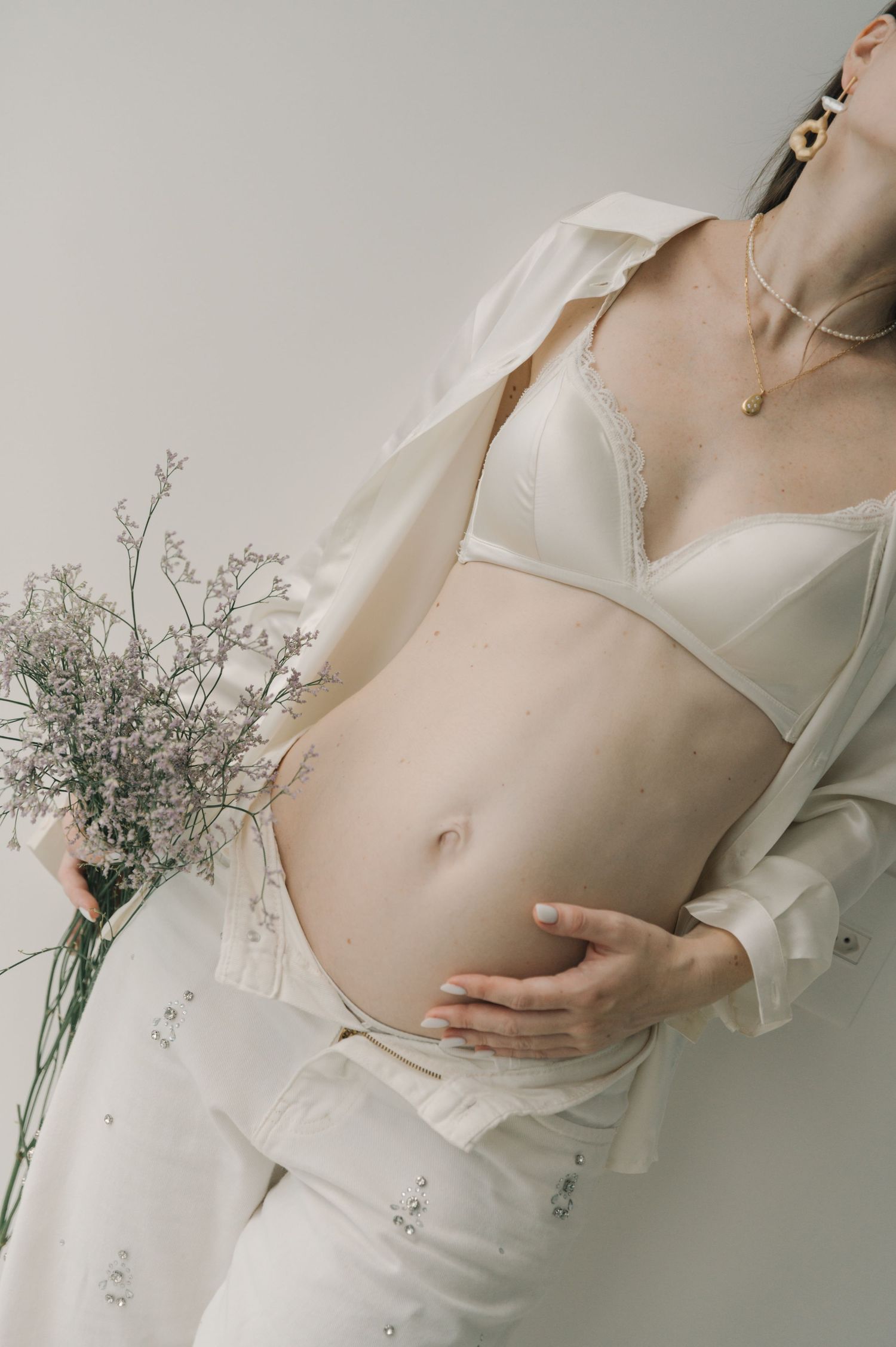 Ethereal maternity photo with dried flowers and white lingerie against neutral background.