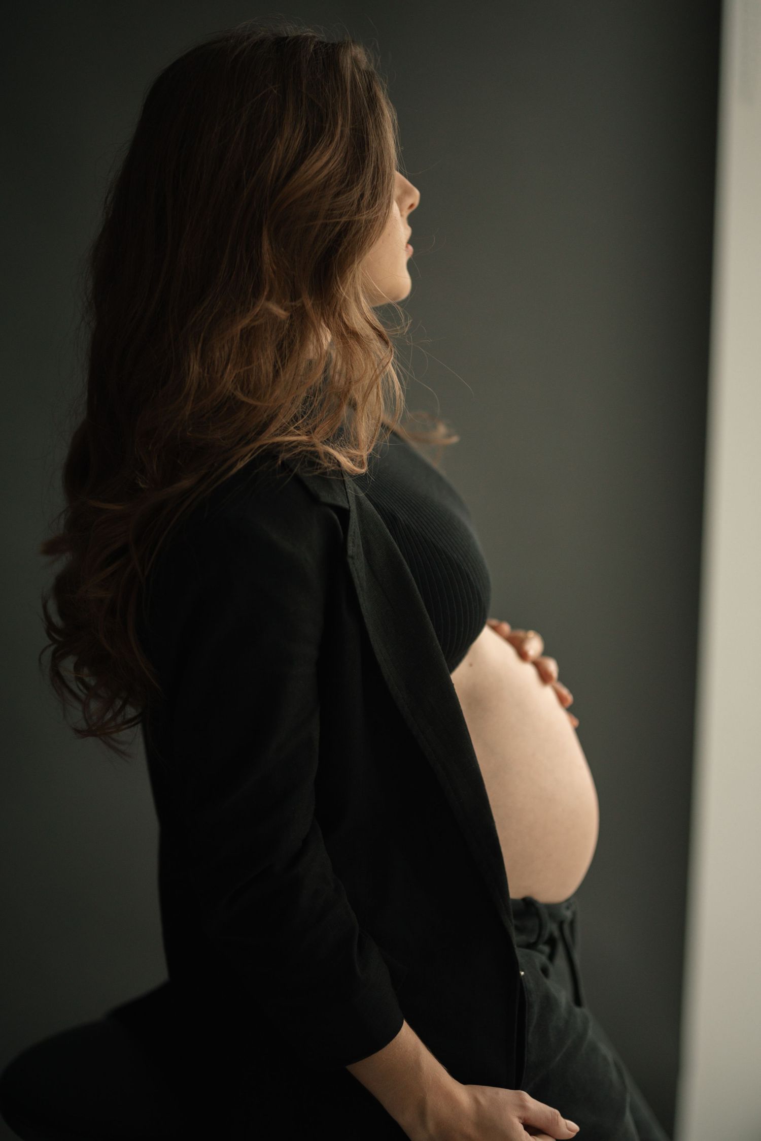 Moody side-view maternity portrait with woman in black dress cradling belly against dark wall.