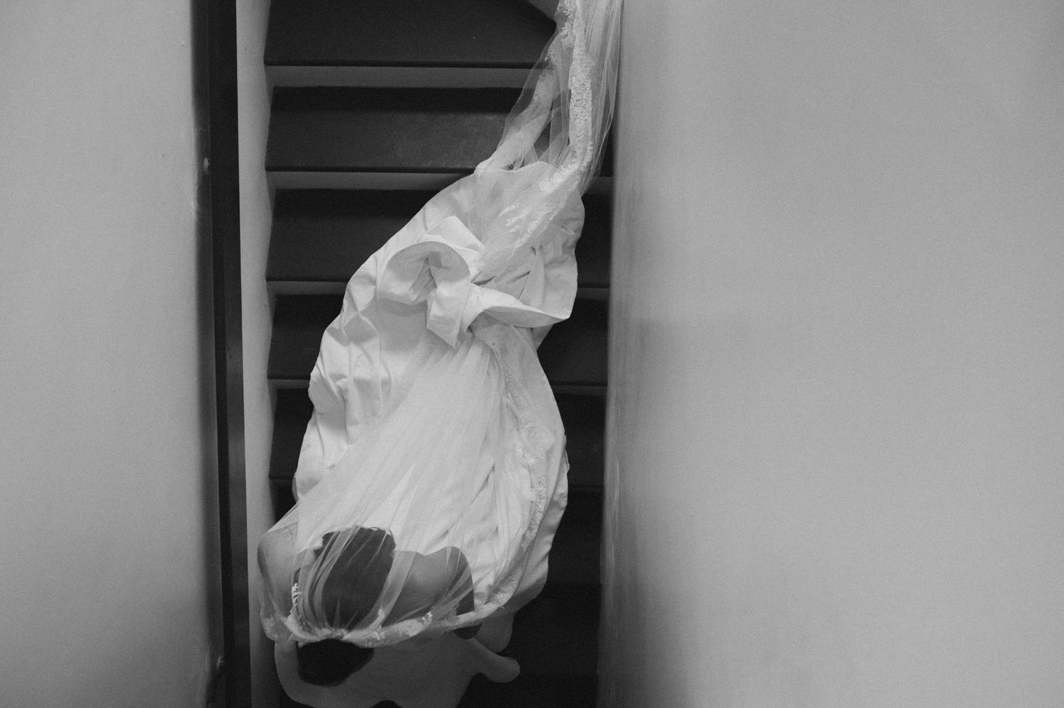 Bride in wedding dress and veil navigating a narrow staircase in black and white photo.