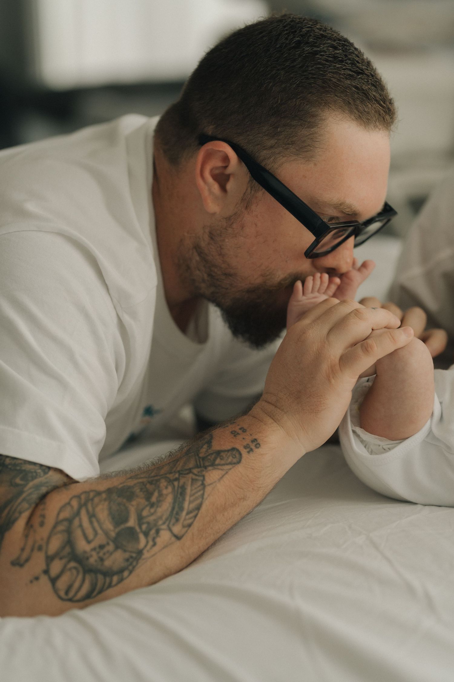 Person with tattooed arm lying on white bedding while covering face showing signs of distress or exhaustion.