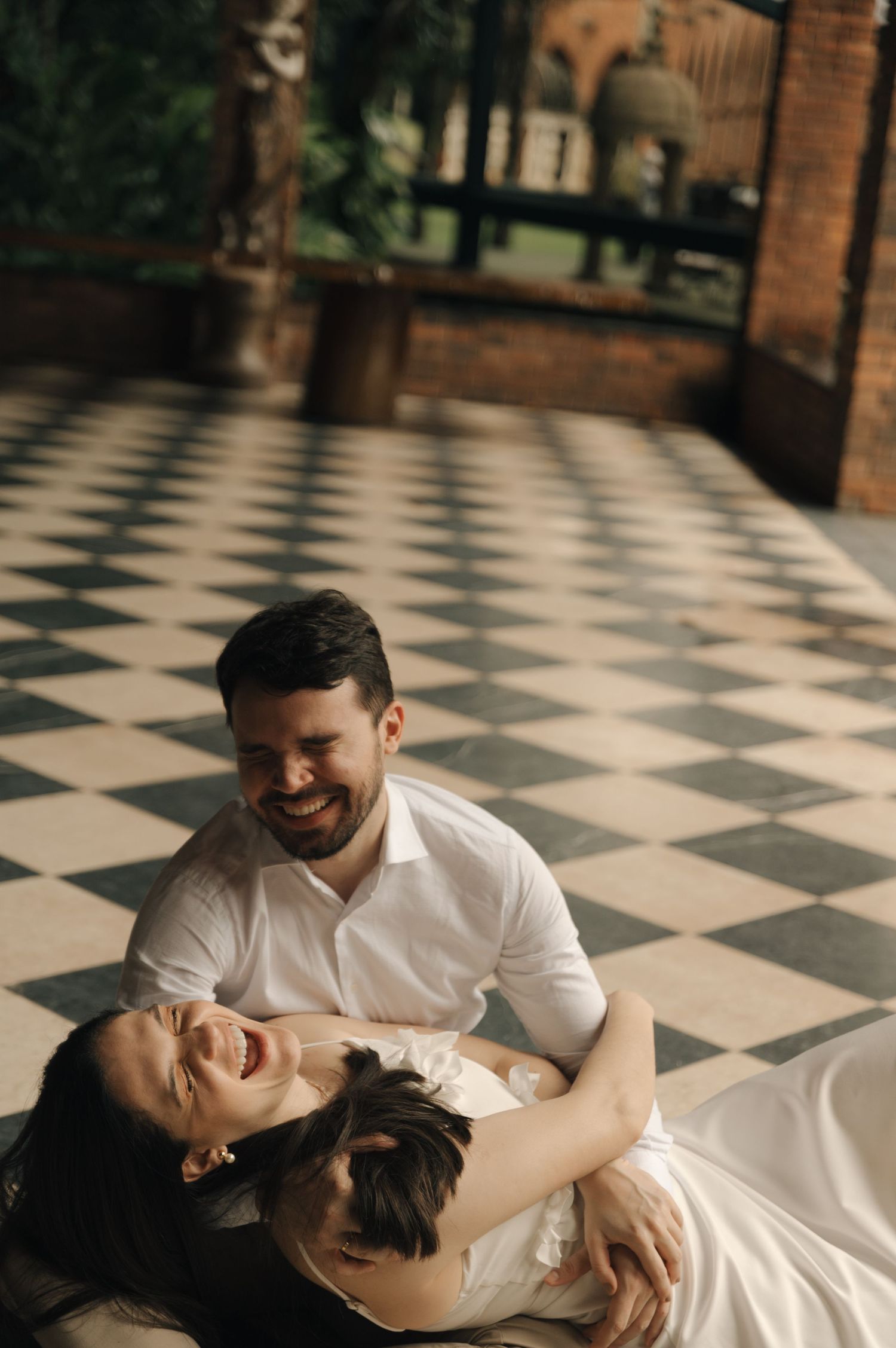 A couple in white outfits laughs together while lying on a black and white checkered floor.
