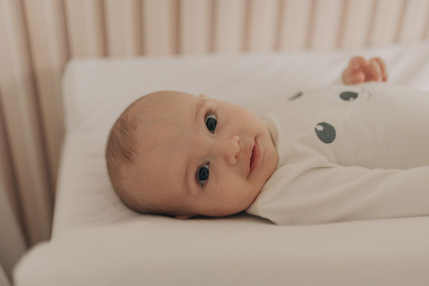Baby lying on white bedding looking directly at camera with bright eyes.