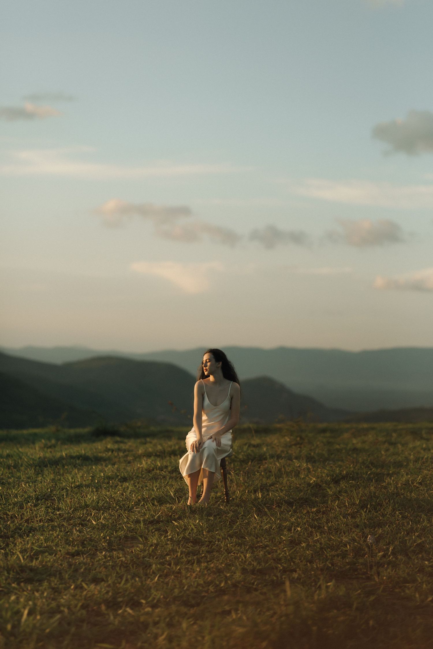 A person in a flowy white dress stands on a hilltop against a sunset sky and mountain landscape.