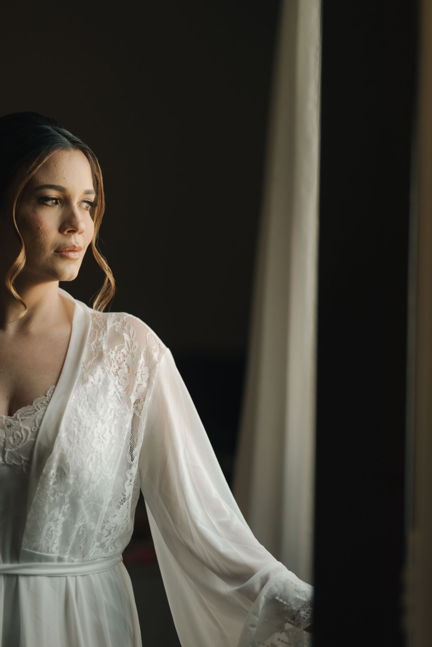 A bride in a white lace robe poses by a window in soft natural light.