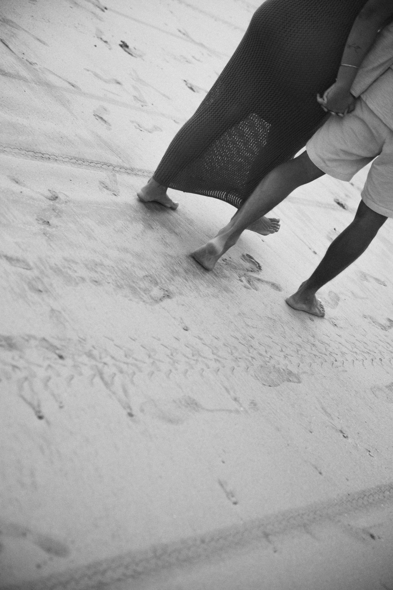 Black and white image of barefoot couple walking on beach sand leaving footprints.