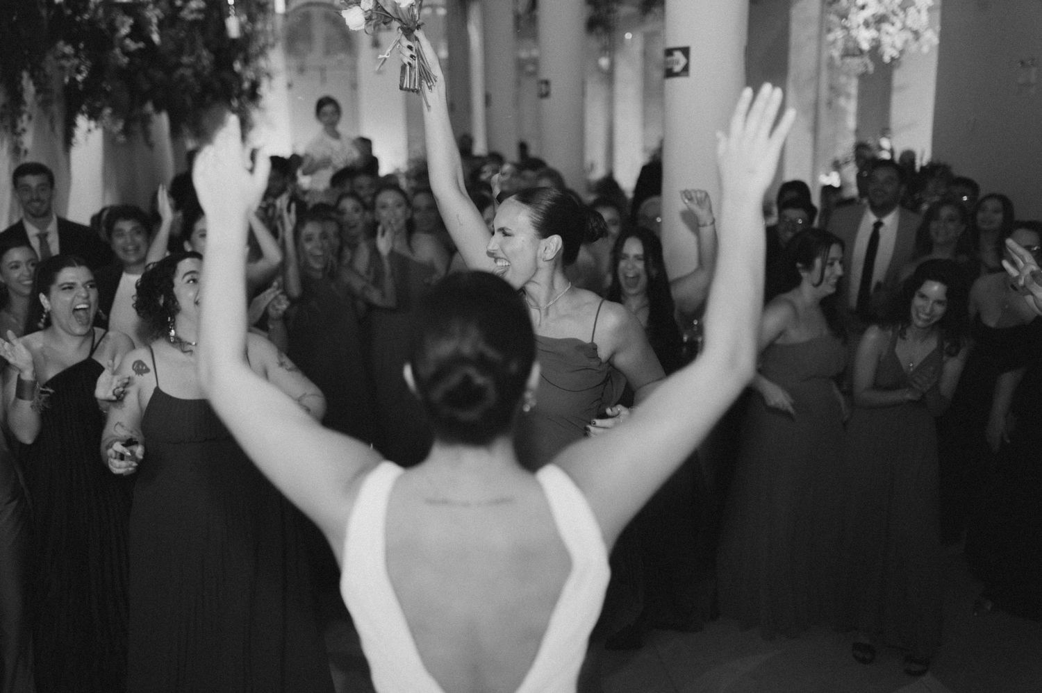 Black and white photo of a crowded dance floor at a wedding reception with raised hands in celebration.