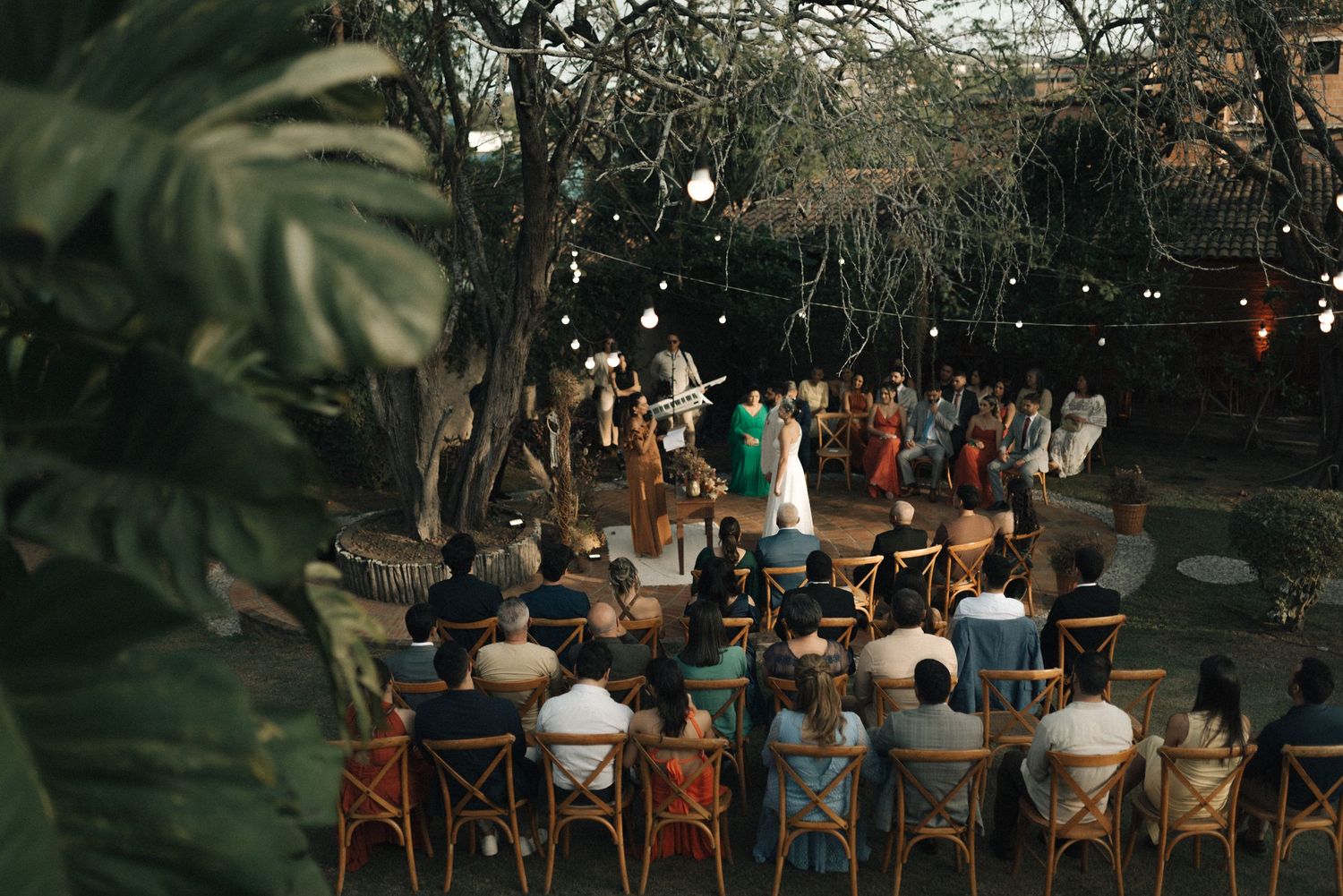 An outdoor wedding ceremony under string lights with guests seated in wooden chairs at dusk.