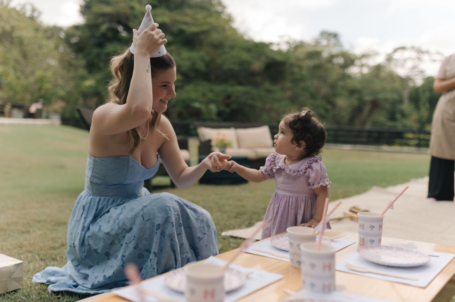 A picnic scene in a park with a parent and child playing together on a blanket with food and drinks spread out.