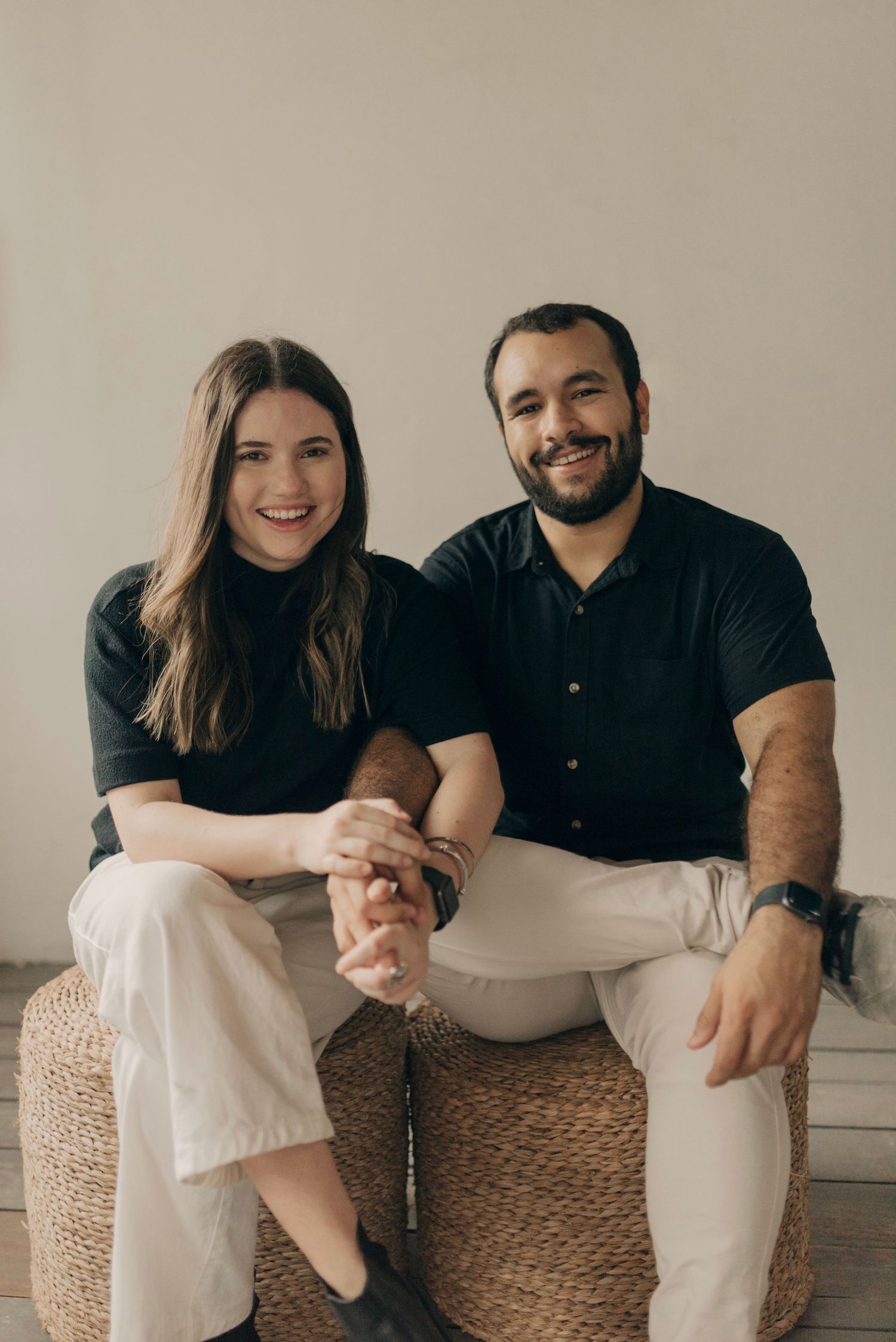 Casual portrait of couple sitting together on a woven ottoman in neutral tones.