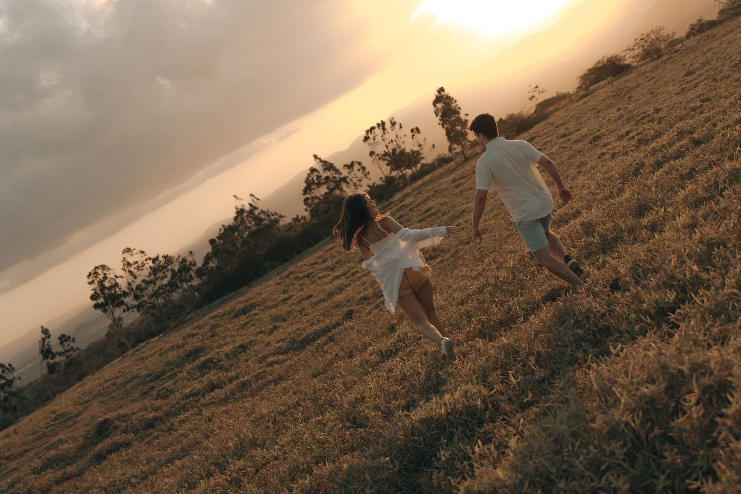 A couple runs playfully on a hillside during golden hour sunset.