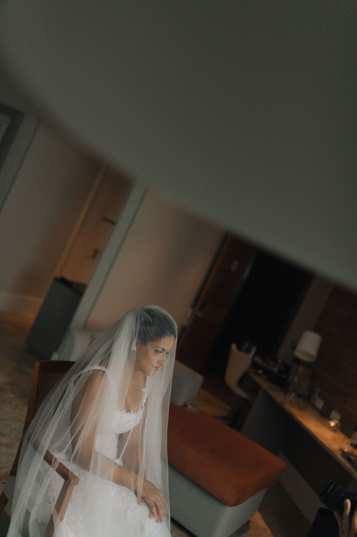 A bride in a white wedding dress and flowing veil prepares in a dimly lit room.