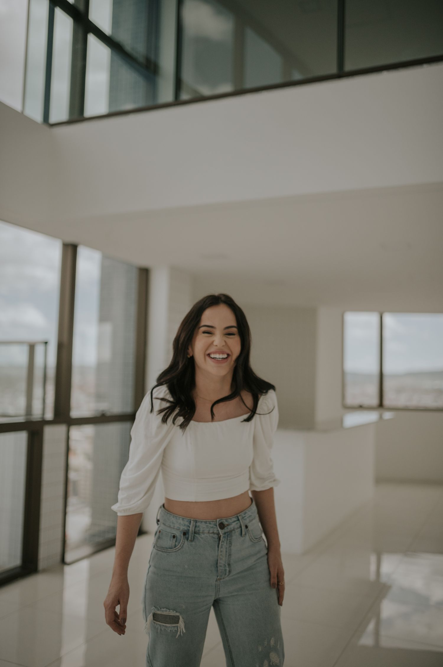 A person wearing a white crop top and distressed jeans poses in a bright modern interior space.