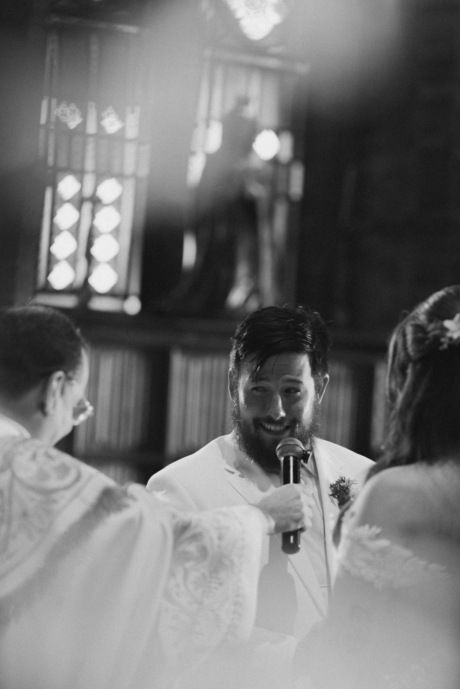 A black and white photograph of a wedding reception scene with guests in formal attire and decorative lighting.