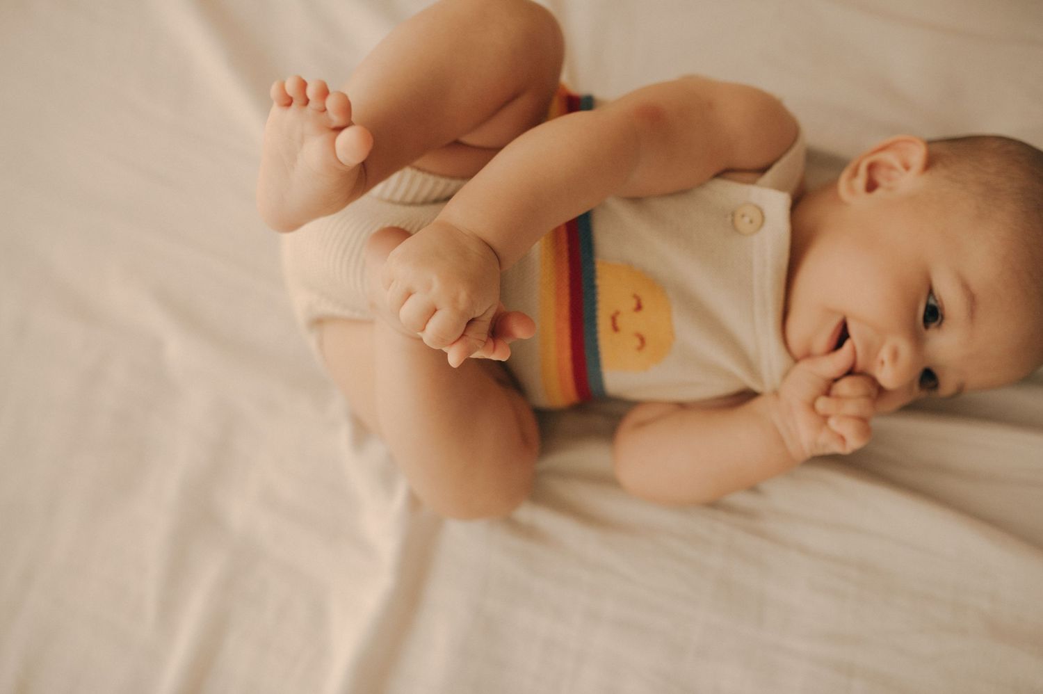Baby wearing rainbow onesie lying on white sheets playing with feet.