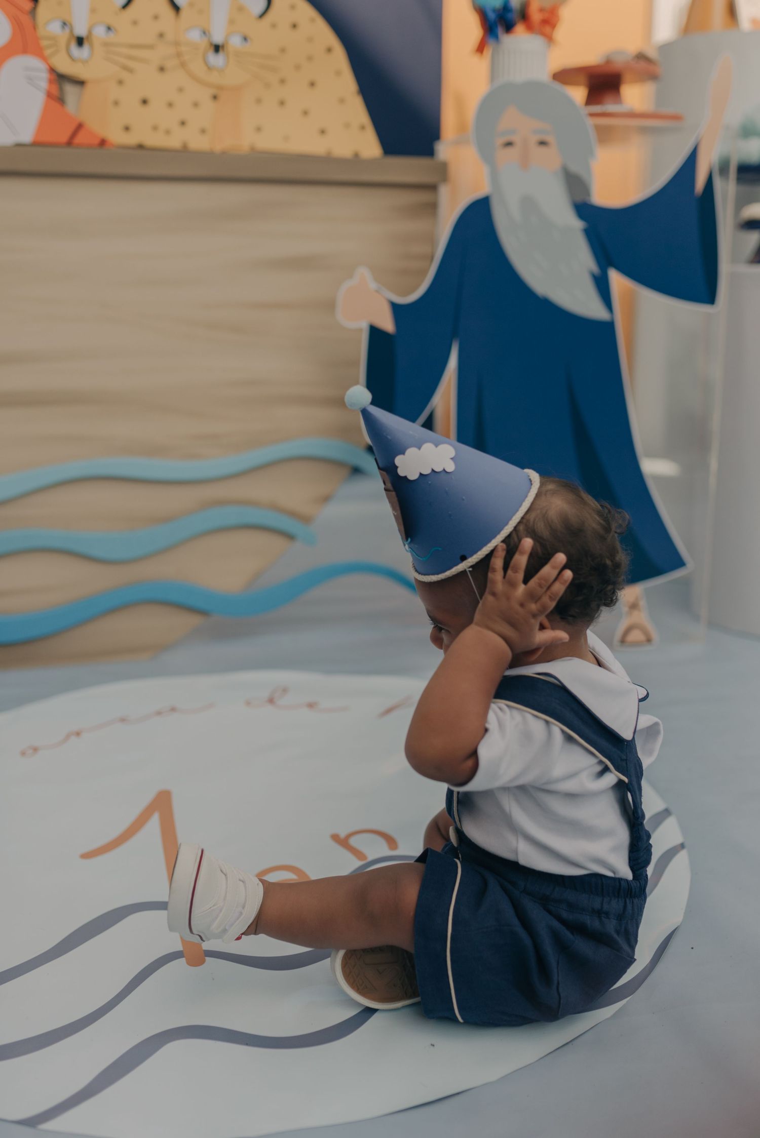 Child wearing party hat sitting near decorative wall mural with whimsical characters.