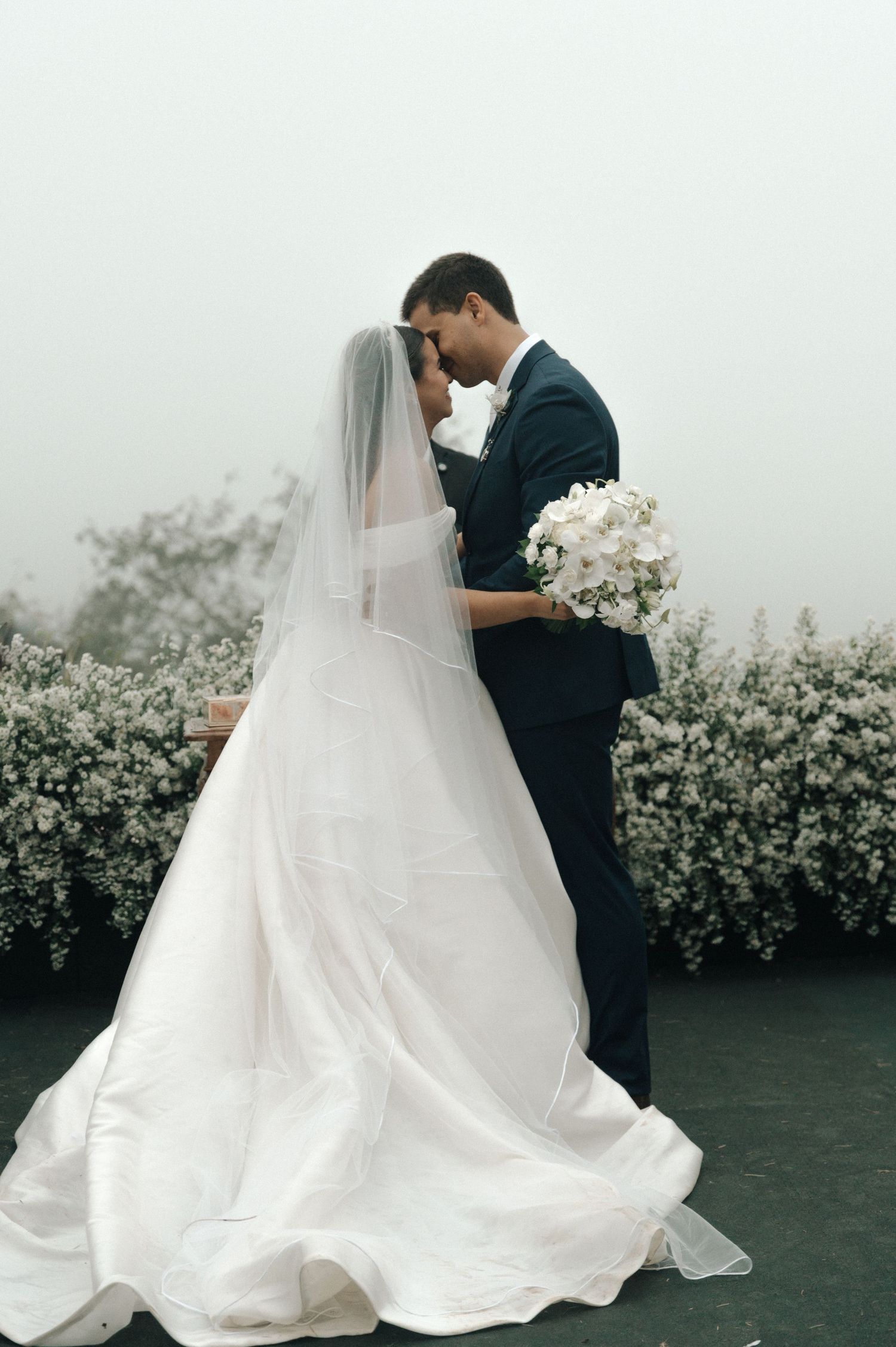 A romantic wedding couple embraces outdoors with flowing white bridal gown and veil against a backdrop of white flowers.